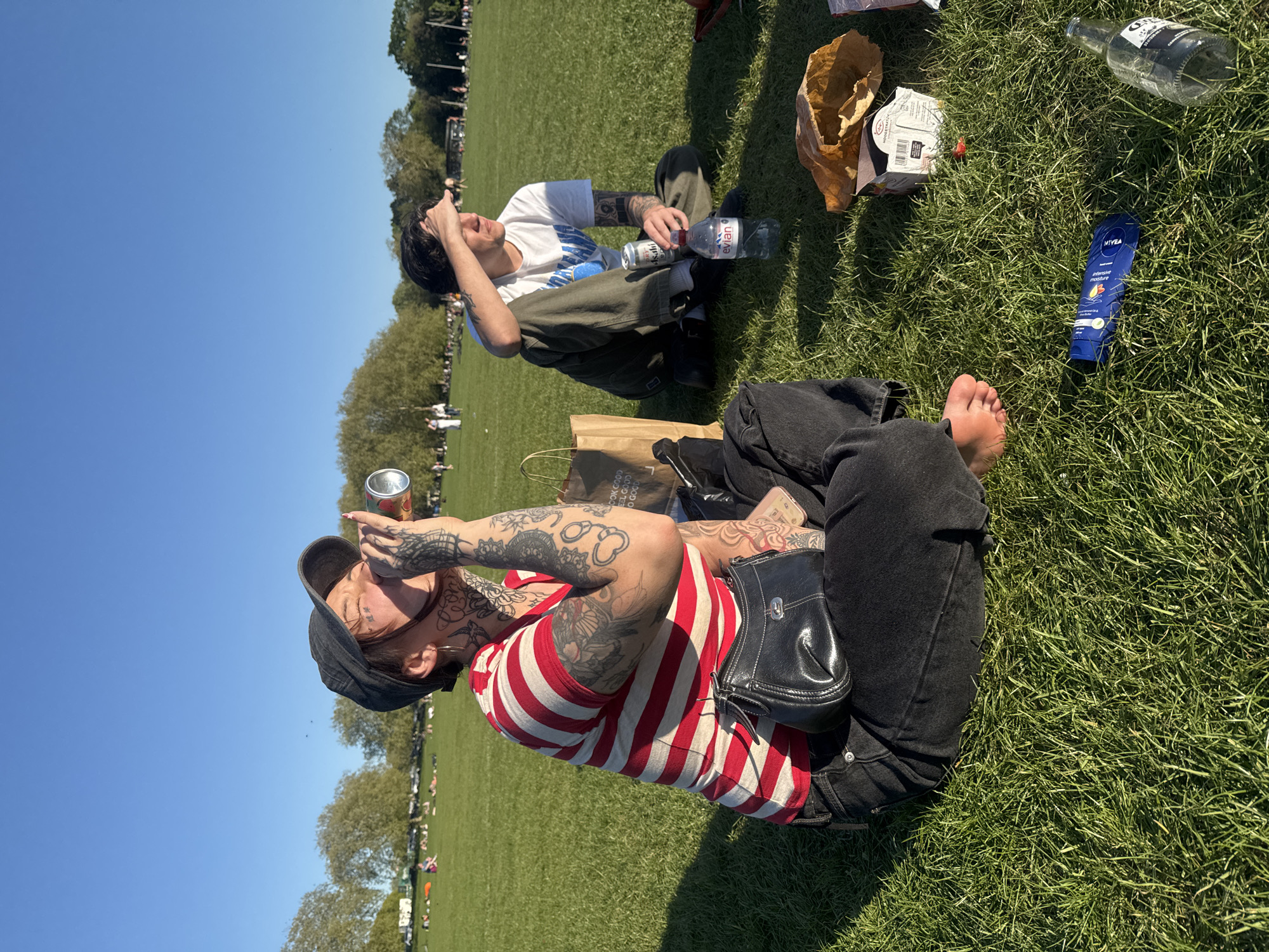 The image depicts two individuals relaxing on a grassy area, likely a park. A person in the foreground, wearing a red and white striped shirt, black jeans, and a black hat, is drinking from a can. Their arms are heavily tattooed. A small black purse is resting on their lap. The second person is in the background, wearing a white t-shirt and green pants, and holding their head. In the vicinity of the two people, there are various items scattered on the grass, including a glass bottle, a plastic bottle, a Nivea lotion tube, and paper bags. The setting appears to be a public park with other people present. The blue sky suggests it is a clear day. The location is Hyde Park, London.
