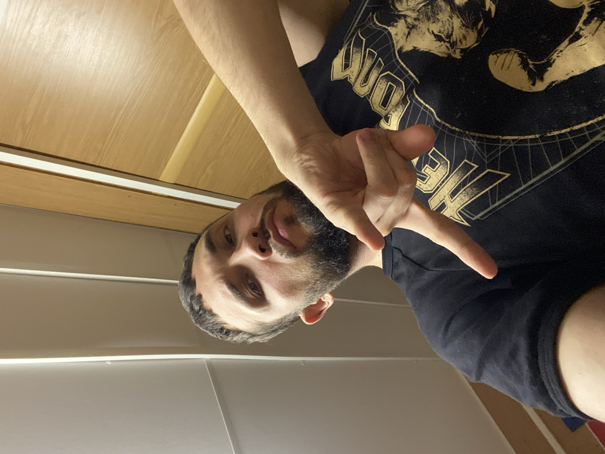 A bearded man is posing for a selfie indoors. He is wearing a black t-shirt with a golden design. The background shows blinds and a wooden cabinet. There is no verifiable evidence to confirm the image's origin in Koskolovo, Russia.