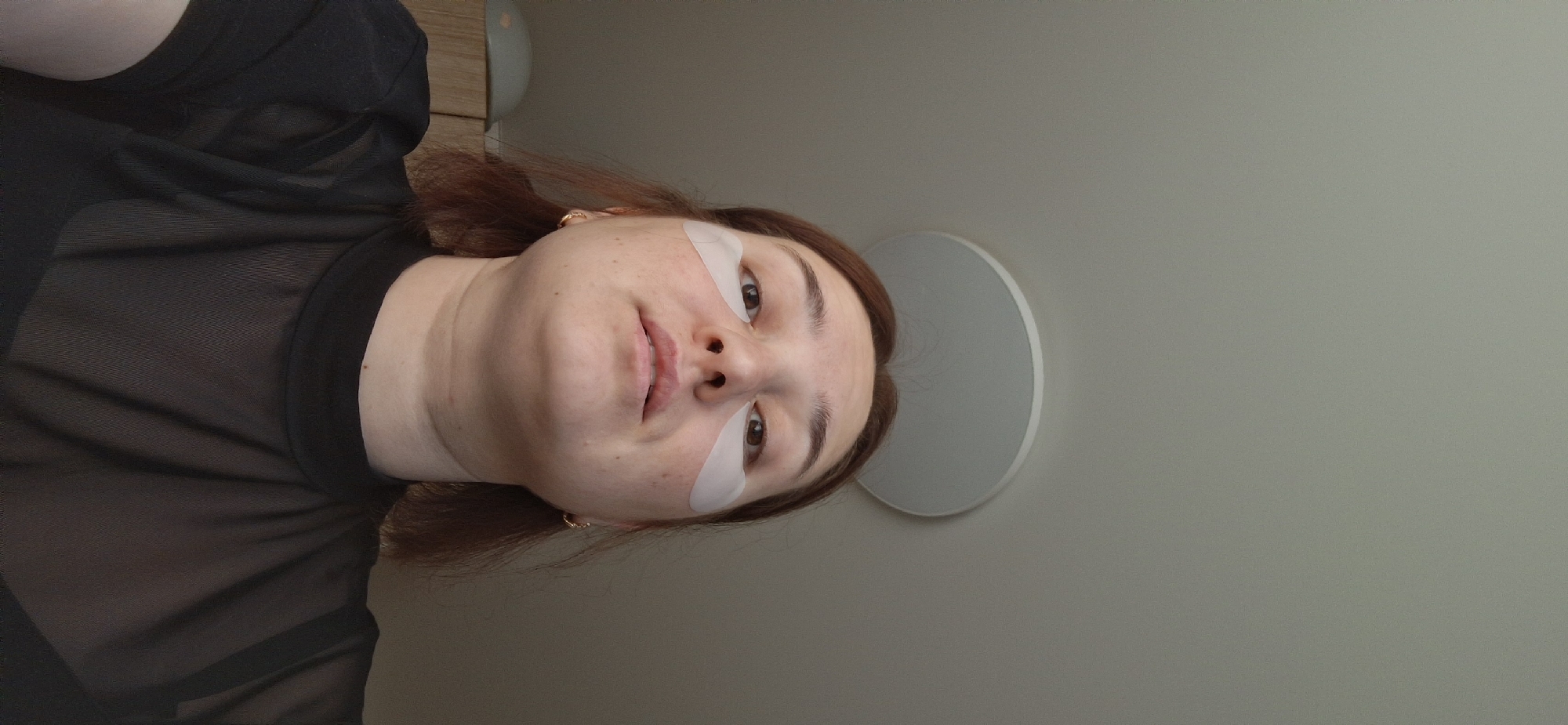 A woman is depicted in a selfie shot at an upward angle. She has light skin, brown hair, and is wearing what appears to be under-eye patches. She is wearing a black, sheer top. The background consists of a plain wall with a circular light fixture. The location cannot be determined to be Chaikovsky, Russia with the given information.