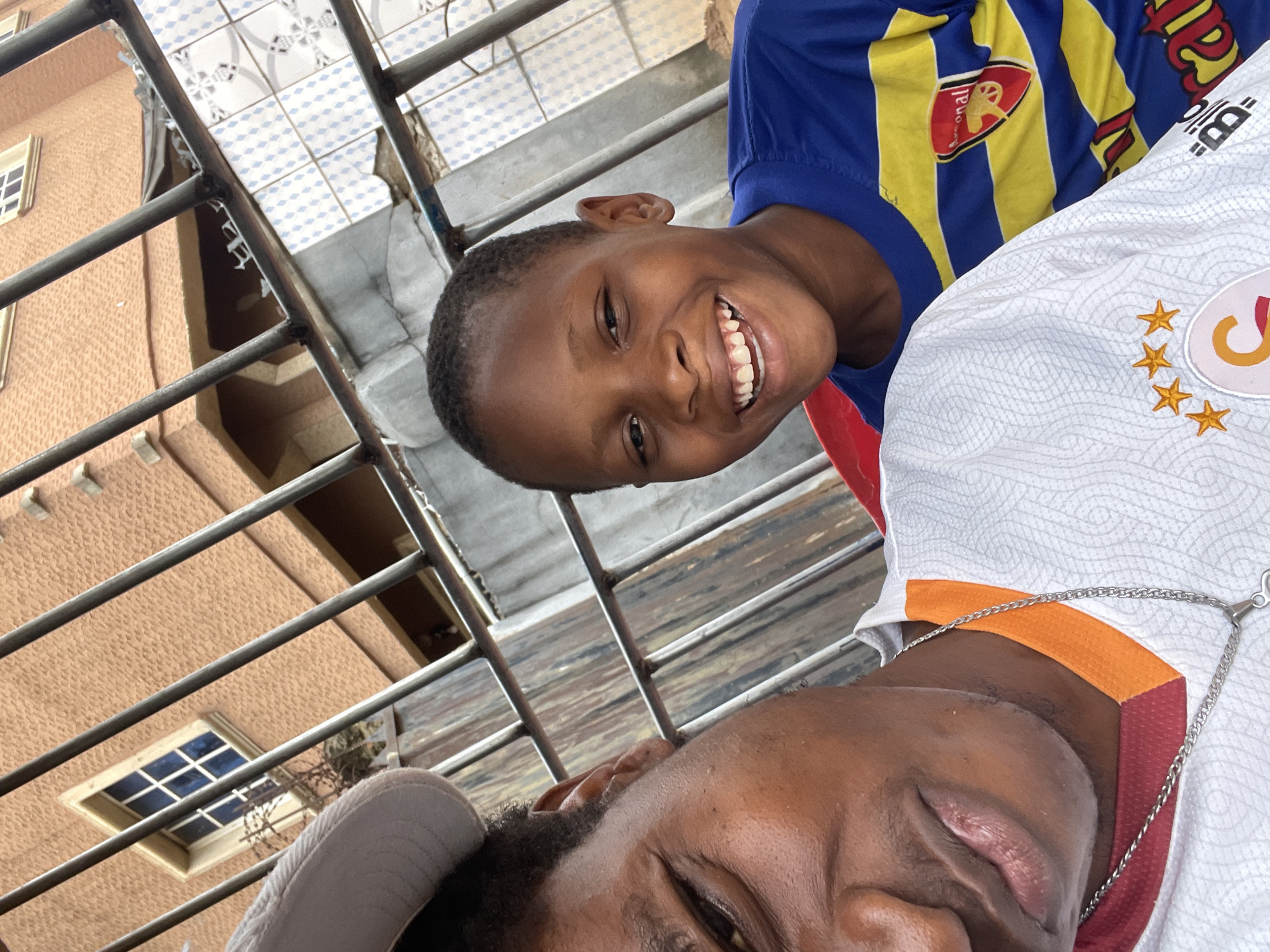 A photograph taken in Onitsha, Nigeria, depicts two individuals, one a child and the other an adult, posing closely together. The child, wearing a blue and yellow Arsenal jersey, smiles broadly at the camera. The adult, wearing a white Galatasaray jersey with a gold star emblem, is partially visible in the foreground. They are positioned behind a metal railing overlooking a residential area. A building with an orange facade and blue-framed windows is partially visible in the background. The ground below the railing is paved with tiled squares.