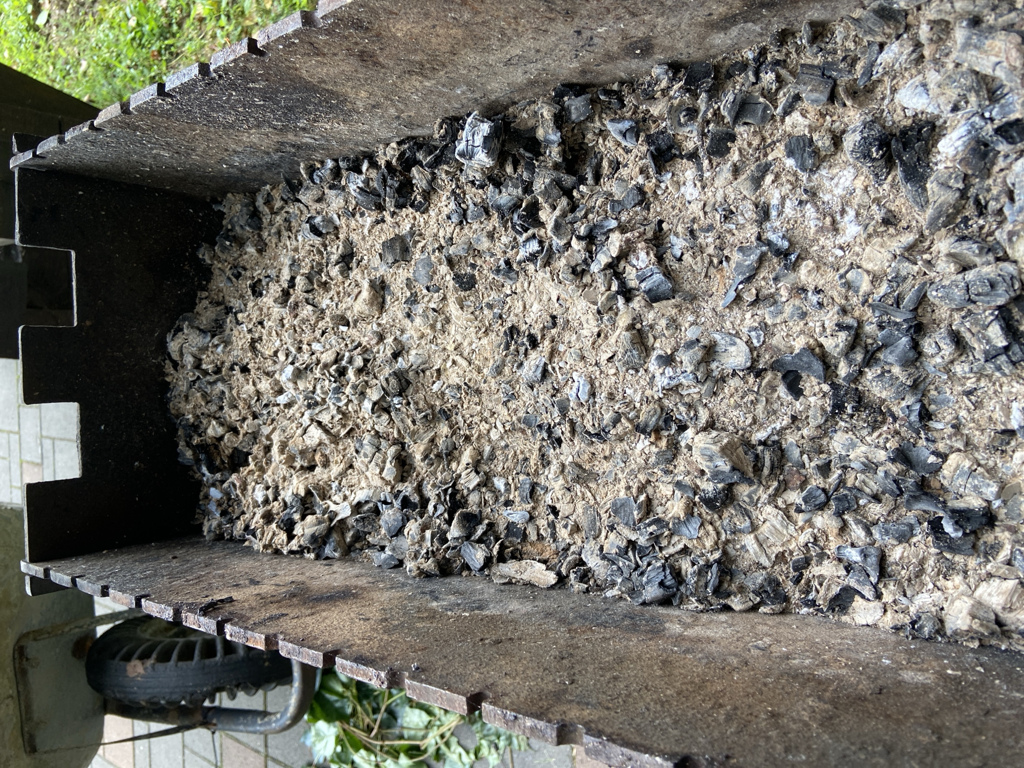 The image shows a close-up view of the inside of a metal barbecue grill filled with ash and charcoal. The grill appears to be old and weathered, with rust visible on the metal. The ash is a light gray color, with numerous dark gray to black pieces of charcoal scattered throughout. The charcoal pieces vary in size and shape, some appearing as large chunks and others as smaller fragments. The grill is situated outdoors, with glimpses of green grass and possibly paving stones in the background, suggesting a yard or garden setting. No people or specific actions are depicted in the image. The location of Зимородок Днп, Russia, is not visually discernible from the image itself.
