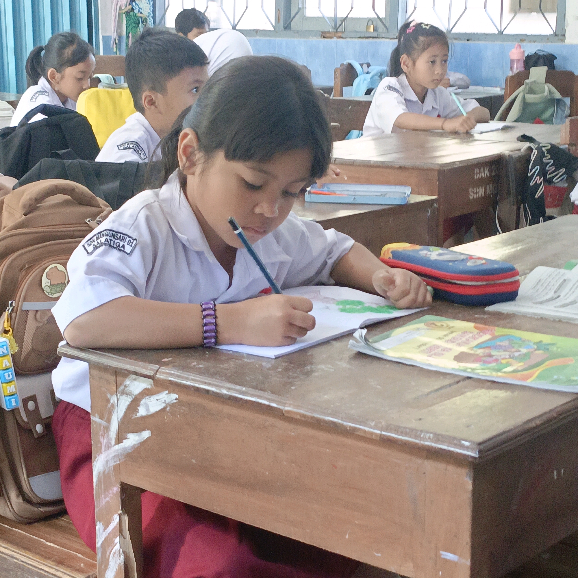 In a classroom setting in Salatiga, Indonesia, a young Indonesian girl in a white school uniform with a red skirt is seated at a wooden desk, diligently writing in a notebook with a pencil. She wears a purple beaded bracelet on her left wrist. Next to her is a brown backpack with a yellow tag attached, displaying the letters "NAUM" in yellow and blue blocks. Another girl, also in a white school uniform, is visible in the background to the left. The desks are made of wood, with visible inscriptions on the front, including "DAK 29 SON MG". The environment appears to be a typical classroom with other students and desks. The image captures a moment of focused activity during a school day.