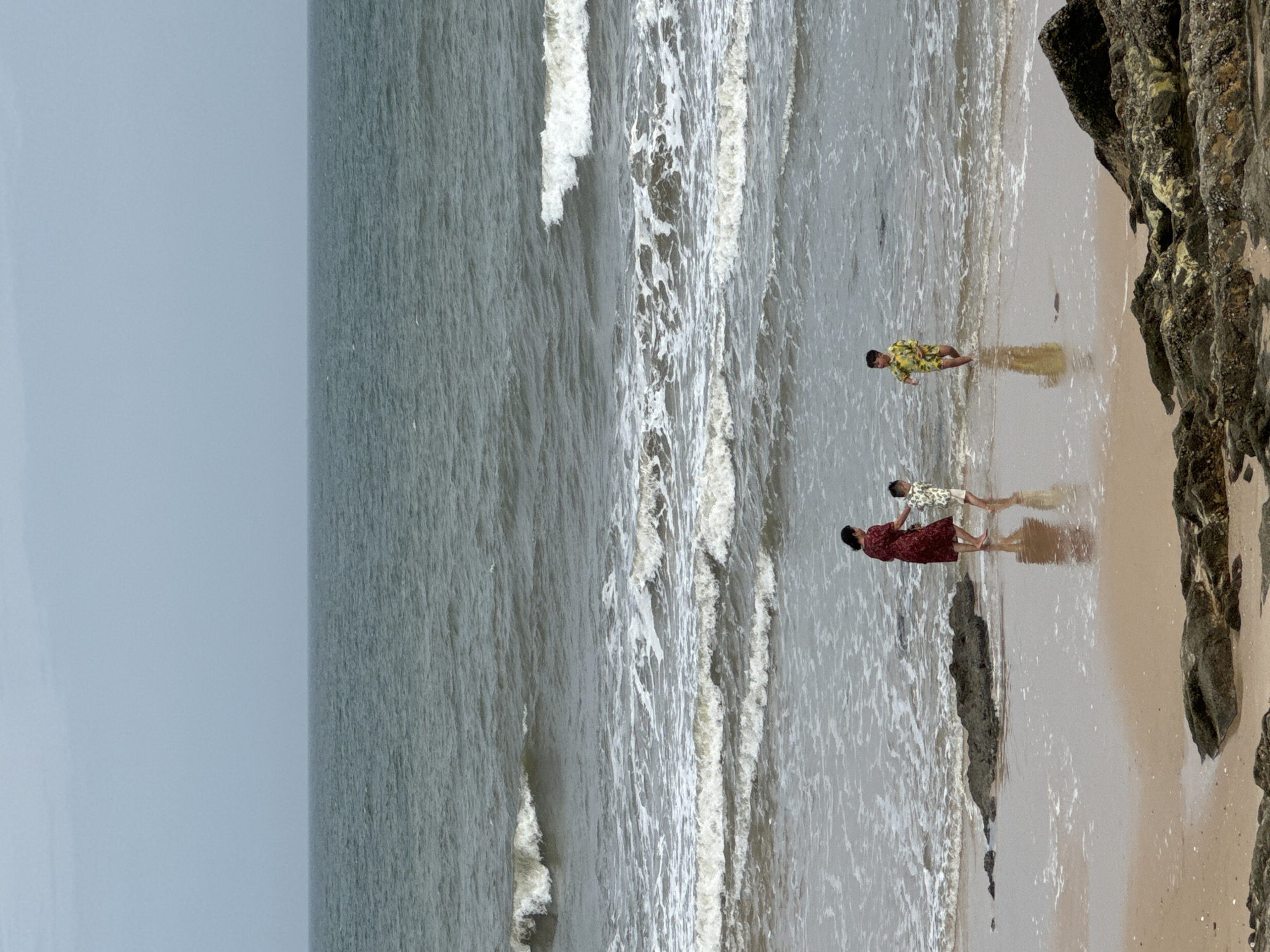 The image shows a family on the beach in Mũi Né, Vietnam. A woman in a red dress and two young boys in patterned swim trunks are wading in the shallow water near the shore. Waves are breaking on the beach, and the sky is overcast. Rocks are scattered on the sand and in the water. The scene depicts a casual outing at the beach with family members enjoying the water.
