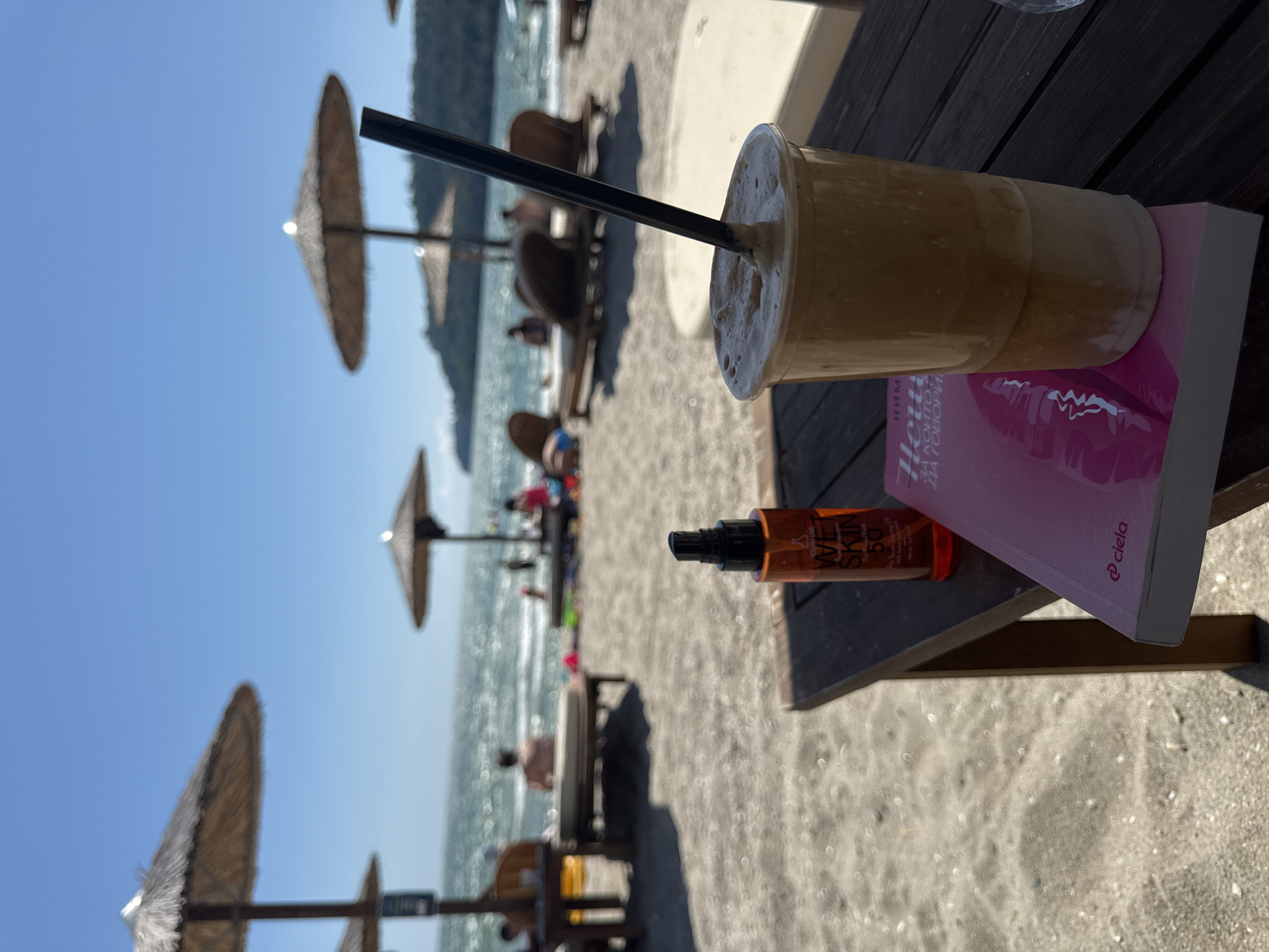The image depicts a beach scene on a sunny day. In the foreground, a dark wooden table holds a clear plastic cup filled with a frothy, light brown beverage with a black straw, placed on top of a pink book. To the left of the cup, a small orange bottle of sunscreen is standing upright. The table is positioned on a sandy beach. In the mid-ground, several thatched umbrellas are visible, providing shade over lounge chairs. Some people are seated or lounging on these chairs, and others are in the shallow water of the ocean. The ocean is calm and reflects the bright blue sky, with a distant green, tree-covered hill forming the background. The overall atmosphere suggests a relaxing day at the beach.