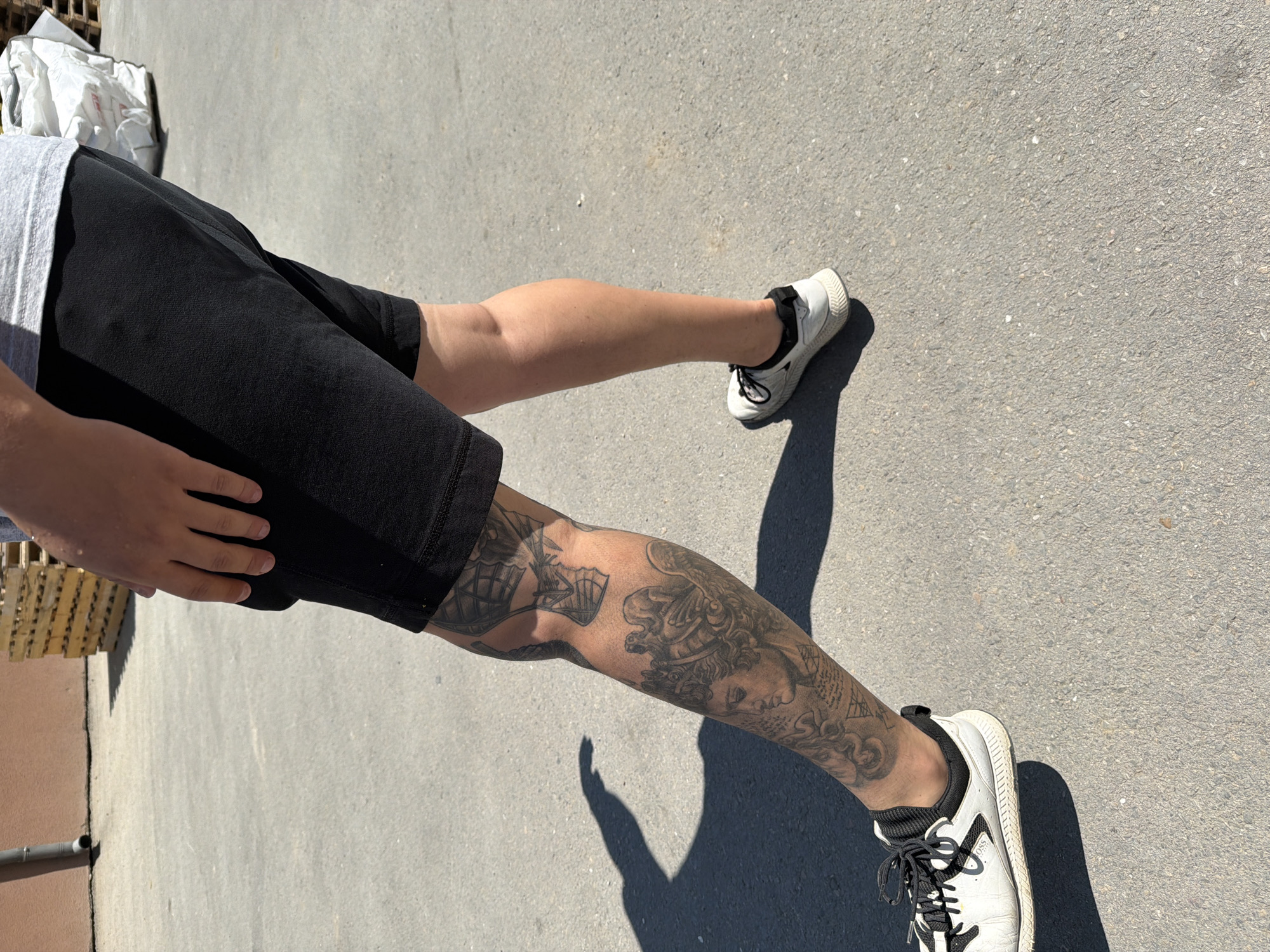 A person's lower legs and feet are visible in this outdoor shot. They are standing on an asphalt surface. The person is wearing black shorts and white sneakers with black laces. The left leg has elaborate tattoos depicting what appear to be mythological figures and possibly a ship. A shadow is cast on the ground by the person's left leg. In the background, there is a building with a downspout, a stack of wooden pallets, and a large white bag. The scene appears to be in Chervena Voda, Bulgaria.