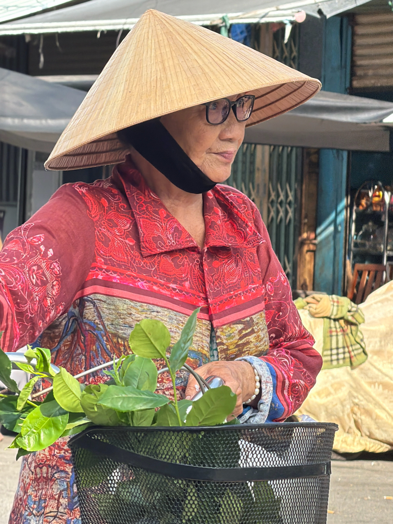 A medium shot shows an elderly Vietnamese woman wearing a conical hat and glasses, with a black face mask pulled down under her chin. She is wearing a red patterned shirt and is looking to her right. In the foreground, a black wire basket holds fresh green leaves, possibly from a plant or herbs. The background depicts a market scene with stalls and goods, suggesting an outdoor urban environment in Nha Trang, Vietnam. The lighting indicates it is daytime, with some shadows suggesting the sun is at an angle. The woman appears to be stationary, perhaps a vendor or a shopper.