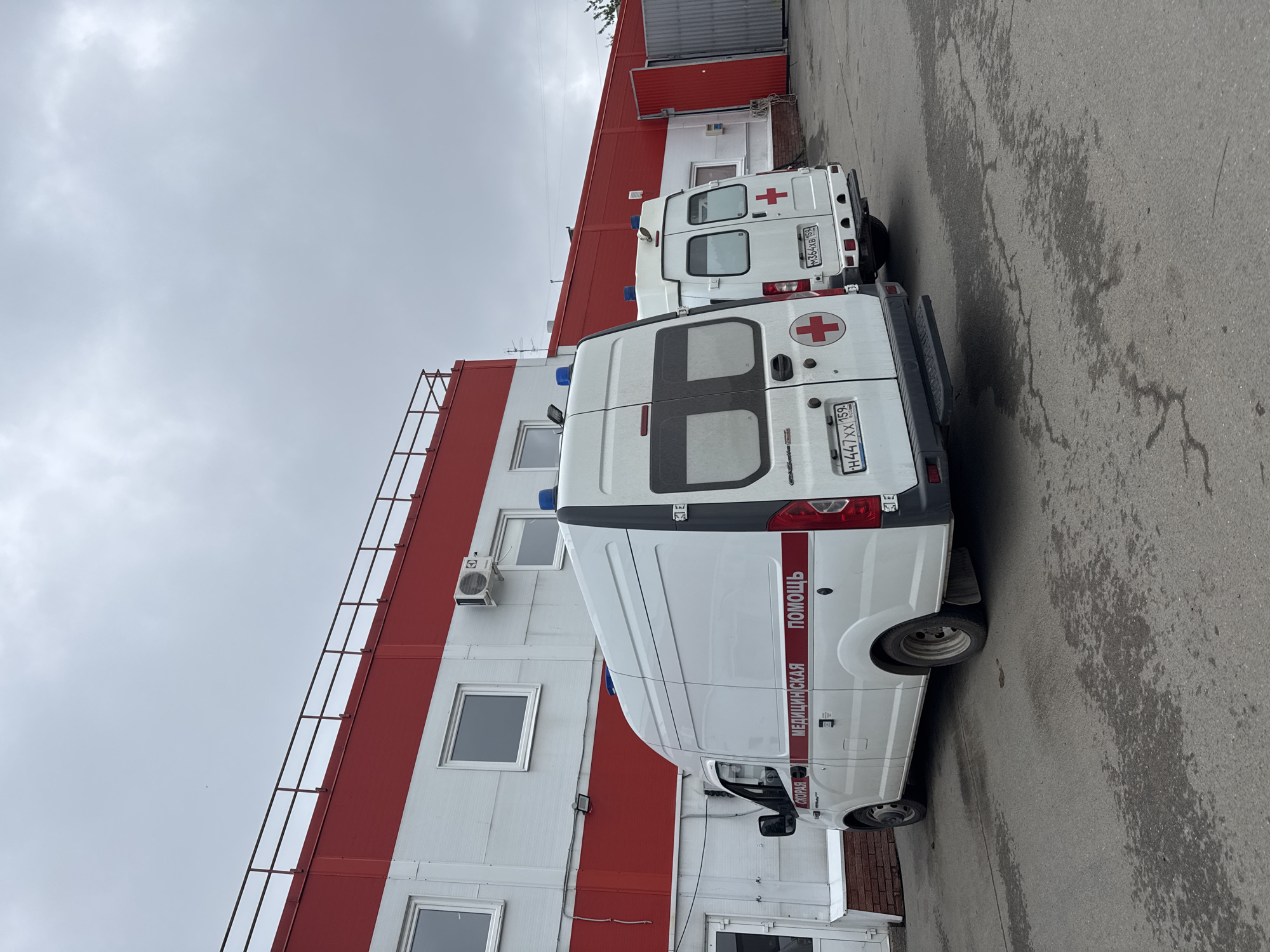 Two white ambulance vans are parked in a paved lot next to a building in Chaikovsky, Russia. The building is white with red stripes and has a ladder running up one side. The vans have red crosses on the back and the words "МЕДИЦИНСКАЯ ПОМОЩЬ СКОРАЯ" ("Medical Assistance Ambulance") written on the sides in Russian. The license plates of the vans are visible. The sky is overcast.