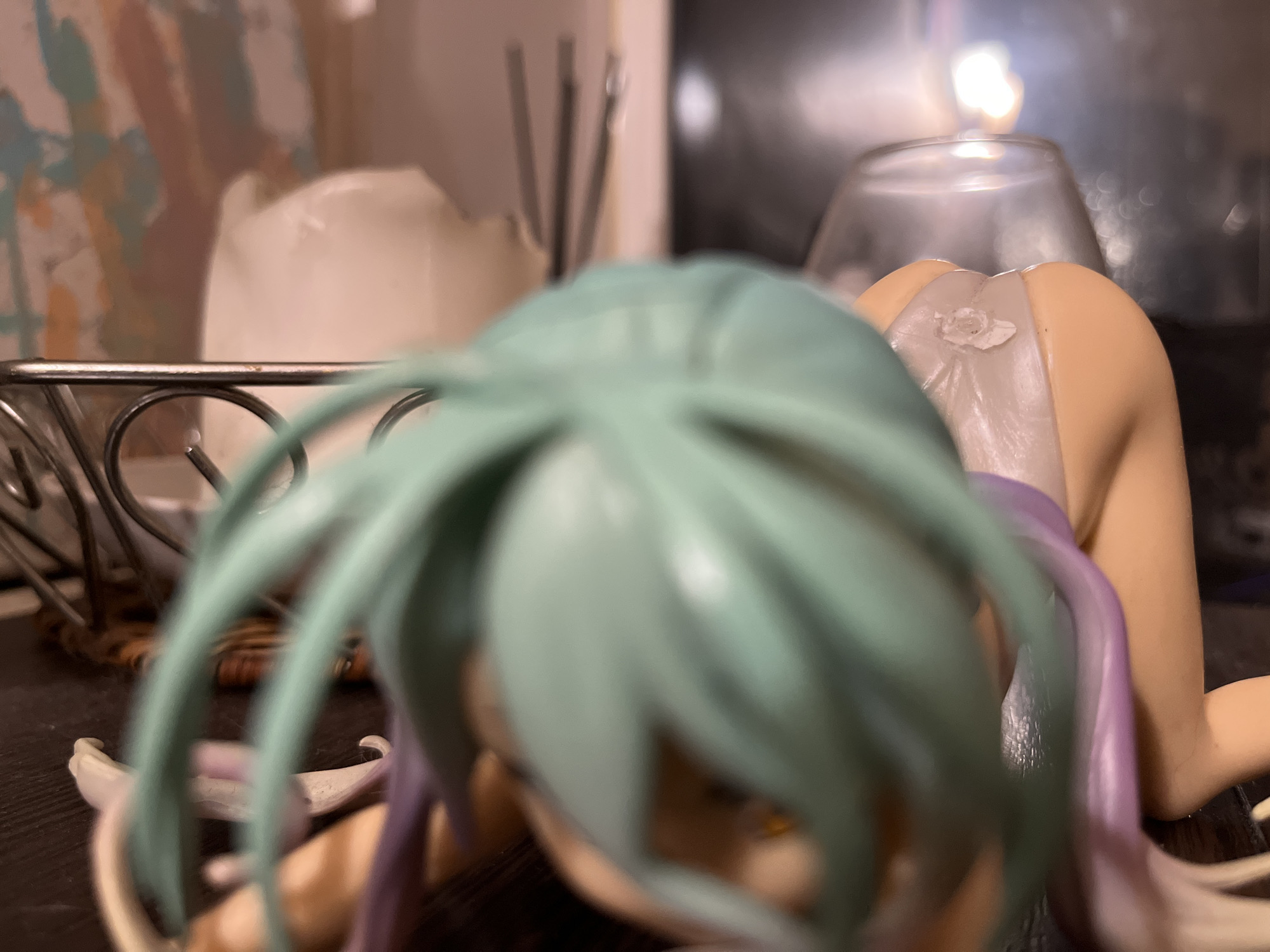 A close-up, shallow depth-of-field, horizontal medium shot shows the posterior of a light-skinned female figurine in a crawling or kneeling pose. The figurine has light purple accents on its body and long, light green hair that drapes over its head and is out of focus. The figurine is wearing light-colored underwear with a small, flower-like detail on the back. The figurine is positioned on a dark brown surface. In the background, a glass object with a bright light emanating from within is visible, along with out-of-focus decorative items and a metallic rack. The scene is indoors and appears to be staged, as indicated by the figurine and its placement.