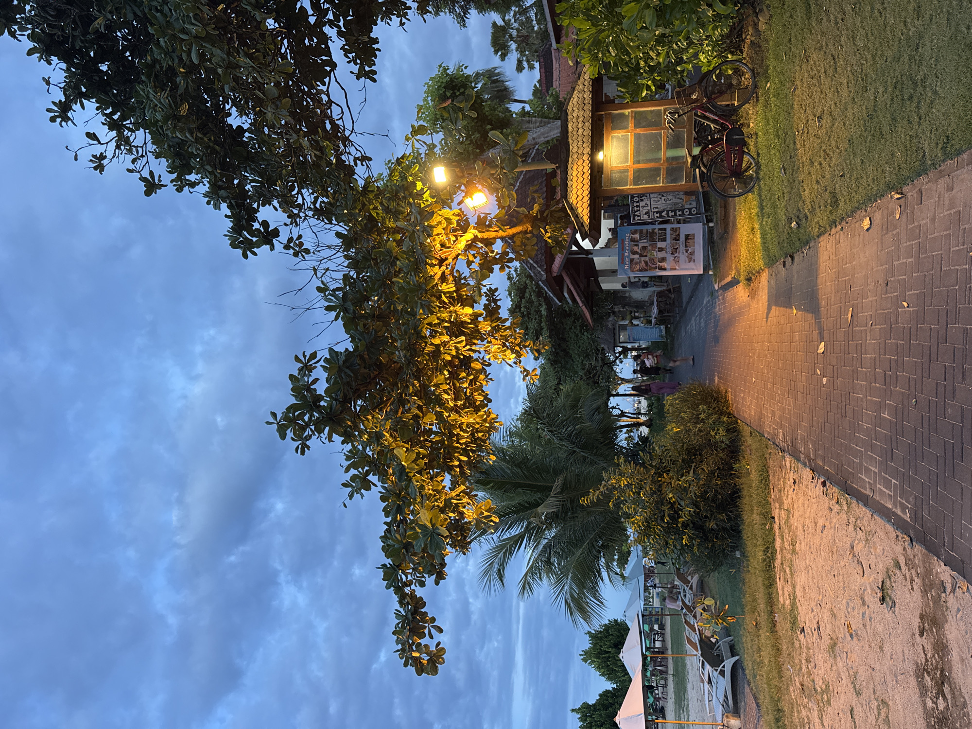 The image shows a pathway at dusk in Kuta Selatan, Indonesia. The pathway is paved with red bricks and is bordered by grass and sand on one side, and a row of small buildings and trees on the other. A small shop with advertisements for hair braiding and tattoos is illuminated by a light, and a bicycle is parked next to it. People can be seen walking along the path. The sky is cloudy and the overall lighting is dim.