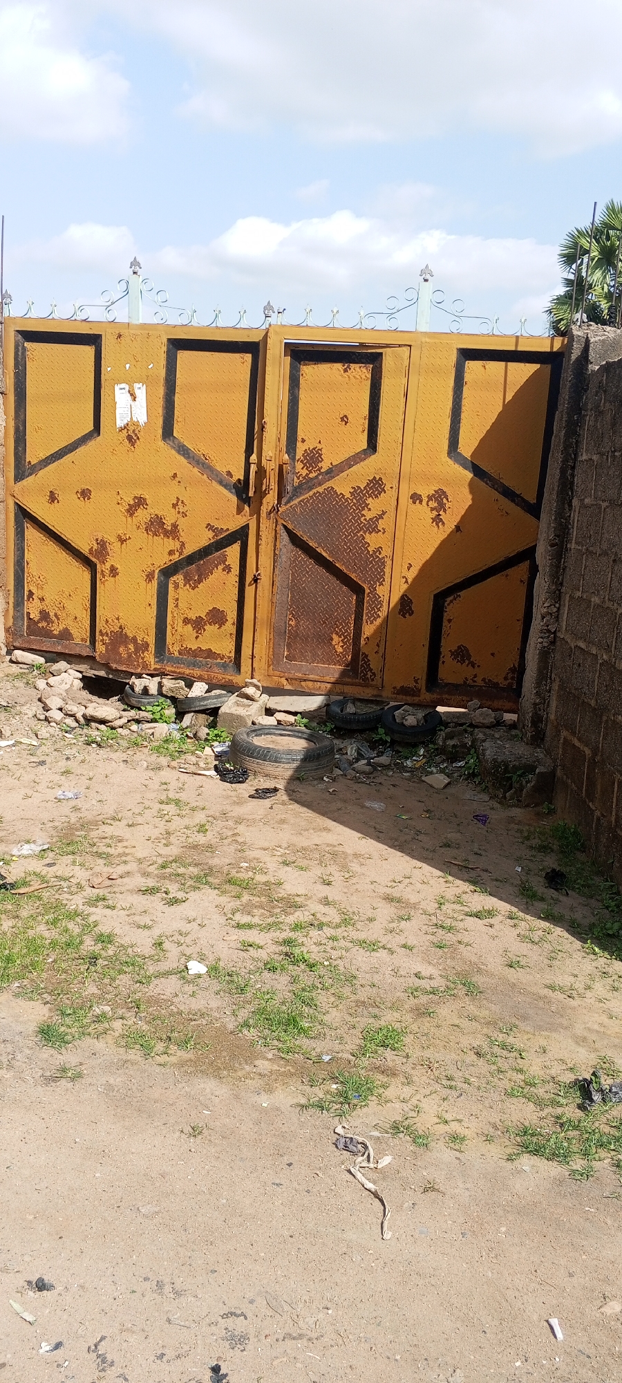 A weathered, yellow metal gate stands in front of a brick wall. The gate is made of two panels, each with a geometric pattern of black outlines filled with textured metal. The yellow paint is peeling and rusty in many places. The gate is adorned with decorative metal scrollwork at the top, some of which is painted a light blue. To the left of the gate, a pile of stones and old tires is visible. The ground in front of the gate is a mixture of dirt and sparse green grass. Scattered on the ground are pieces of trash and a discarded tire. The scene is illuminated by bright sunlight, casting a shadow across the ground from the gate and the wall. The sky above is blue with scattered white clouds. The overall impression is one of an outdoor, somewhat neglected area. There is some unreadable text on a white sticker on the left panel of the gate.