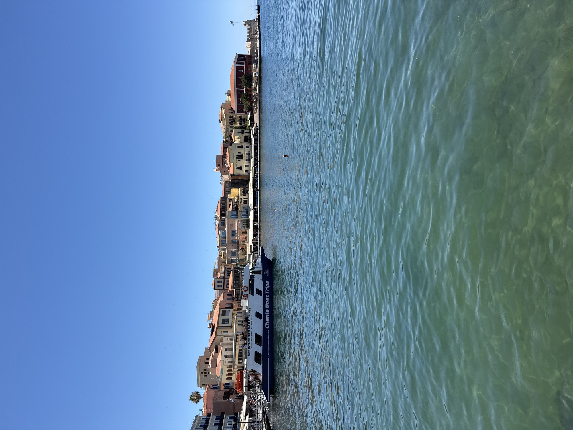 This is a vertical, eye-level, outdoor shot of a bright, sunny day in the city of Chania, Greece. The image captures the clear blue sky and the turquoise water of the harbor. In the foreground, the water shimmers with sunlight reflecting off its surface. A white and blue boat, labeled "KRI-KRI ISLAND DAILY CRUISE" and "Chania Boat Trips", is docked on the left side of the frame. People are visible on the boat and on the adjacent dock, where tables and umbrellas suggest a waterfront dining area. The background features a row of traditional Chania buildings with terracotta roofs, characterized by their colorful facades and Venetian architecture. A lighthouse-like structure with a Greek flag flying is visible on the right side of the harbor, marking the entrance to the port. The overall scene depicts a vibrant and picturesque harbor in Chania.