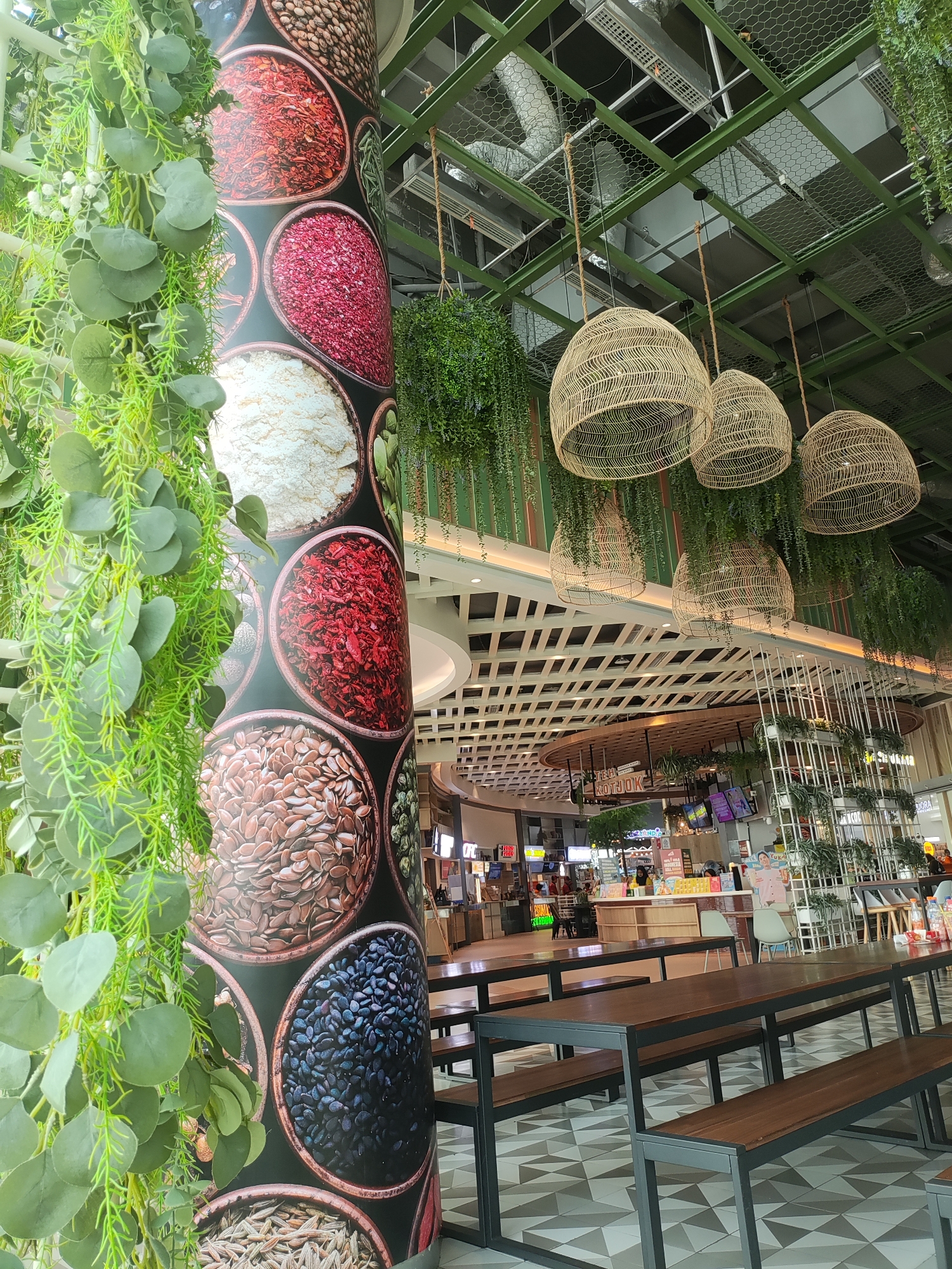 The image depicts the interior of a modern food court, likely located in Lambangjaya, Indonesia. In the foreground, a large cylindrical pillar is adorned with a wrap featuring close-up images of various spices and grains in circular bowls. Hanging plants, including what appears to be eucalyptus leaves and smaller trailing vines, cascade down from the ceiling on the left side of the pillar.
Above, the food court ceiling features a green metal grid structure with hanging rattan light fixtures shaped like inverted baskets. More hanging plants are suspended from the grid. The background reveals the bustling food court environment with multiple food stalls, seating areas with wooden tables and benches, and shoppers. The flooring is a geometric pattern of grey and white tiles. The overall atmosphere is contemporary and vibrant, with a focus on natural elements like plants and woven materials.