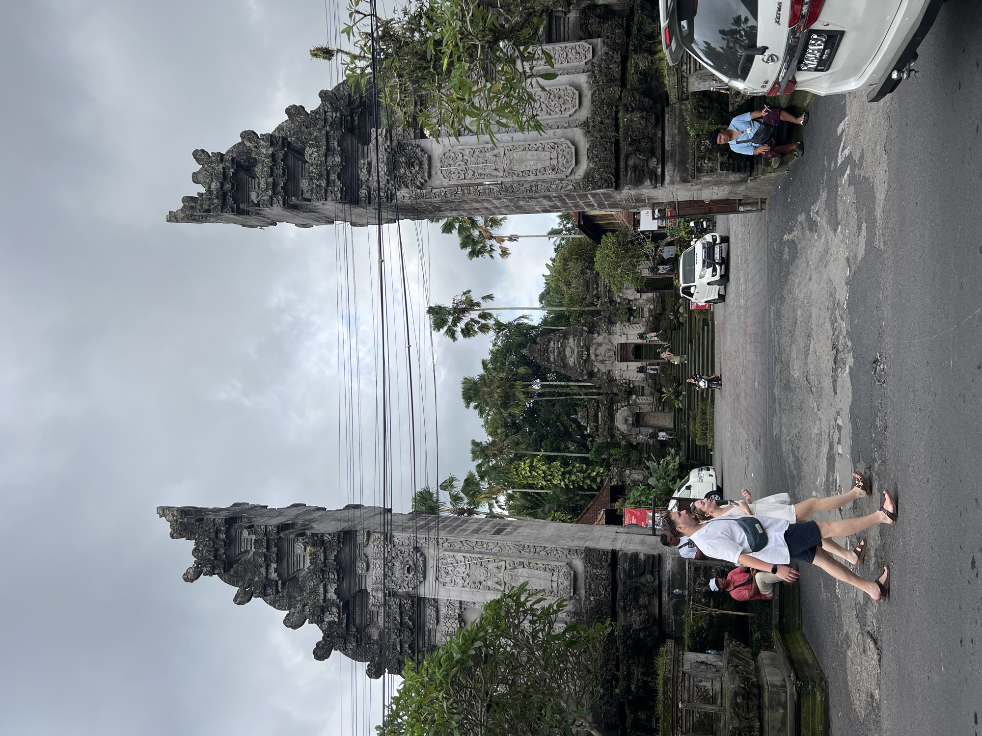 This image depicts a scene in Ubud District, Indonesia, featuring a traditional Balinese split gate (candi bentar). Two elaborately carved, gray stone structures flank a road, creating a grand entrance. Overhead, a network of electrical wires stretches between the gates.
People are present in the image. A couple walks along the right side of the road, with the man wearing a white shirt and dark shorts and the woman in a white dress. Other people can be seen in the background. A man is sitting next to a white car.
A white van is parked further down the road, and a motorcycle drives in the distance. Lush tropical vegetation, including palm trees, fills the space beyond the gate, leading to a set of stairs. The sky is overcast with gray clouds. The pavement is dark and shows signs of wear.
