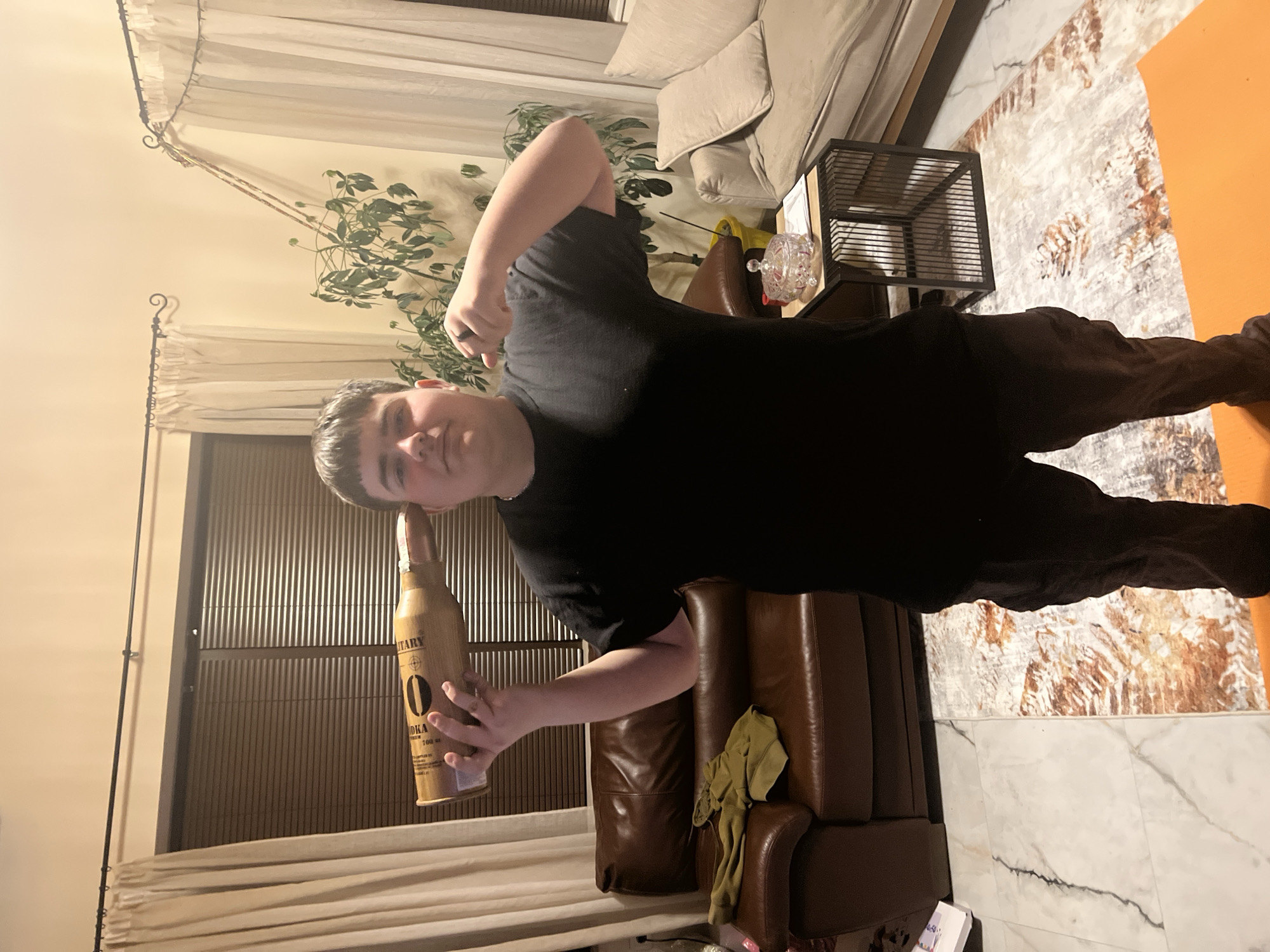 A young male individual stands centrally in a domestic interior located in Berkovitsa, Bulgaria. He is observed wearing a black t-shirt and dark cargo-style pants. His left hand holds a large, elongated, light-brown object, shaped like an artillery shell, with its pointed end positioned near his left temple. Text partially visible on the object includes "VODKA" and "MILITARY." His right hand is clenched into a fist at chest level, displaying a ring on the index finger. He maintains a direct gaze towards the camera with a neutral to slightly smirking facial expression.
The background consists of a living area furnished with a brown leather sofa on the left, draped with a folded olive-green garment. Behind the subject, a tall, leafy houseplant in a yellow pot is visible. To the right, a lighter-colored fabric chaise lounge or sofa section is present, alongside a small, dark-framed side table. On this table rests a clear, lidded glass dish and some papers. Windows fitted with horizontal blinds are partially visible behind light-colored curtains hanging from dark decorative rods. The floor features white marble-patterned tiles and a rectangular area rug with an abstract pattern of browns, oranges, and off-white tones. An orange mat is partially visible beneath the subject's feet. The room is illuminated by warm, artificial overhead lighting.