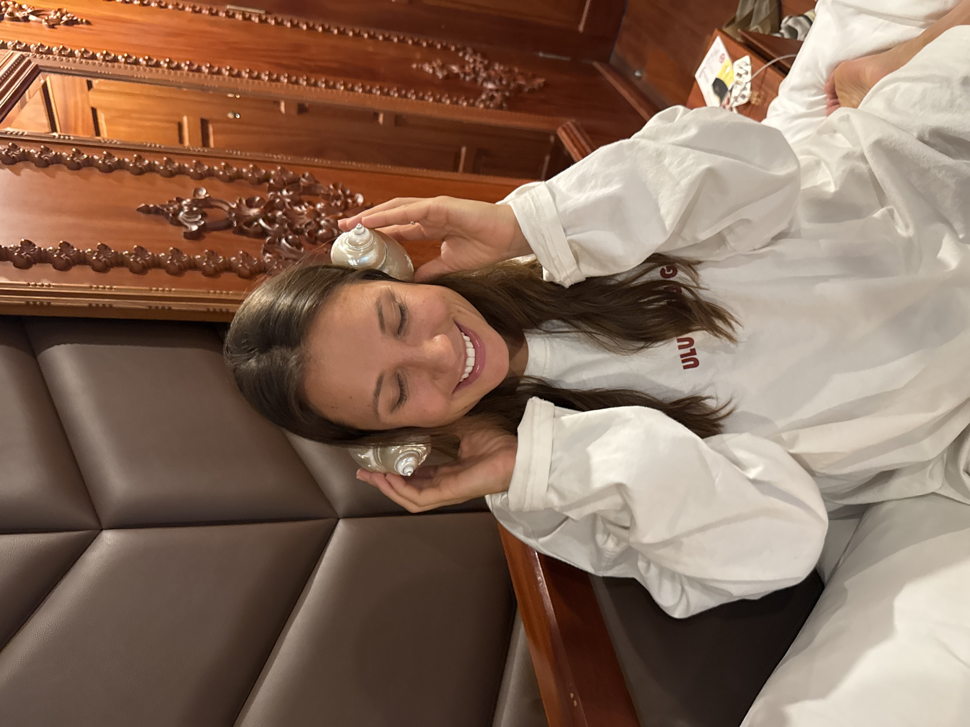 A smiling woman is lying in a bed in what appears to be a hotel room, holding two conch shells near her ears. She is wearing a white long-sleeved shirt. The bed has a brown headboard with a geometric pattern. There is carved wooden trim on the walls of the room. The woman is most likely at InterContinental Danang Sun Peninsula Resort in the city of Đà Nẵng, Vietnam.