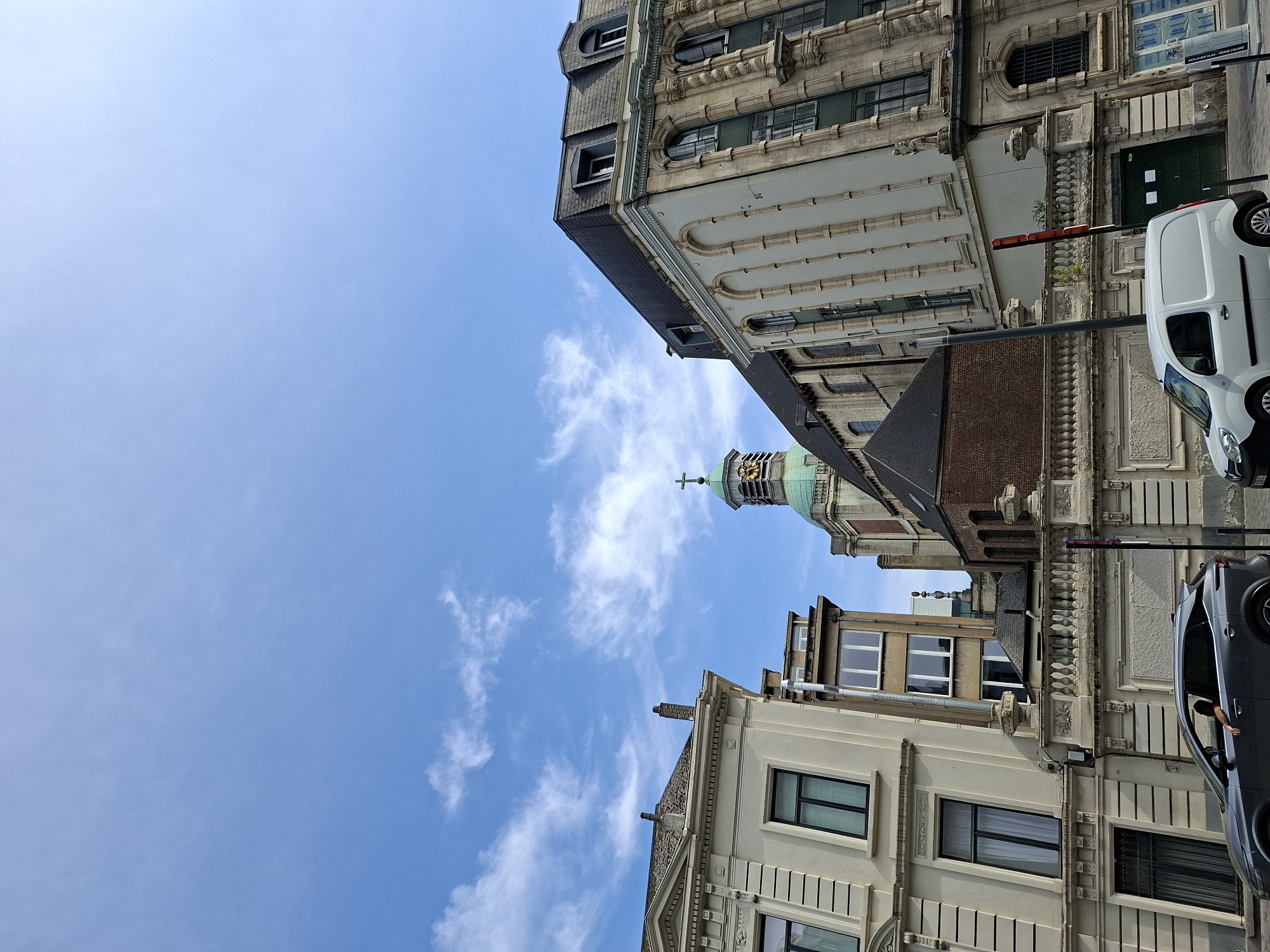 The image shows a street in Brussels, Belgium, on a sunny day with scattered clouds. In the foreground, a grey SUV is parked, and a white van is driving away. A person is visible in the driver's seat of the SUV, with their arm resting on the open window. Behind the vehicles, several buildings are visible, including a church with a green dome and a clock tower, and ornate stone buildings with large windows. The sky is a clear blue with wispy white clouds. The architecture suggests a historical European city.