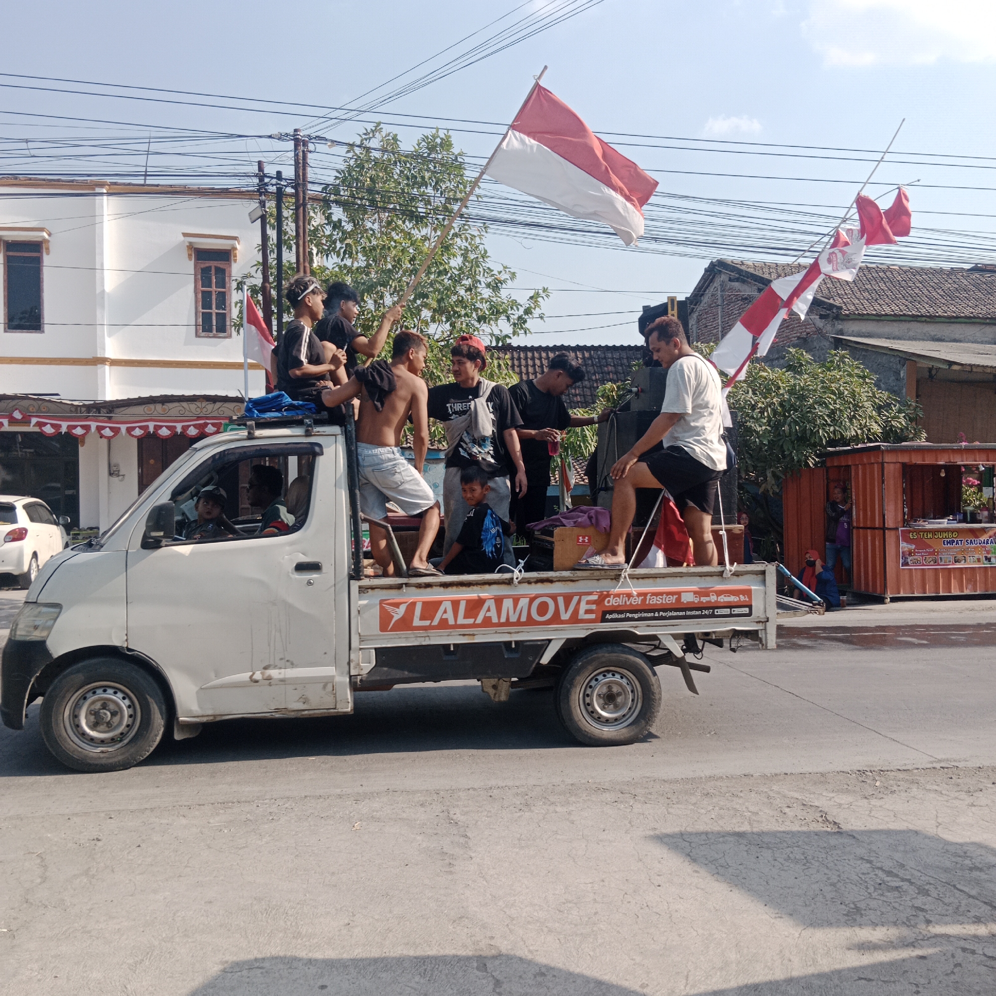This is a medium shot, eye-level, outdoor, daytime photograph taken from a side-view of a white pickup truck driving on a paved road in what appears to be a residential or commercial area of Tegalrejo, Indonesia. The truck is carrying several young men, most of whom are wearing black t-shirts and some are holding red and white Indonesian flags. The flags are displayed prominently, suggesting a patriotic event or celebration. One of the men is standing on top of a speaker system on the truck bed. The truck itself has the "LALAMOVE deliver faster" logo on its side. In the background, there are buildings with white walls and red-tiled roofs, trees, and power lines. A white car is parked on the left side of the road. The sky is clear and blue. The overall scene depicts a group of people celebrating or participating in a procession while traveling in a truck.
