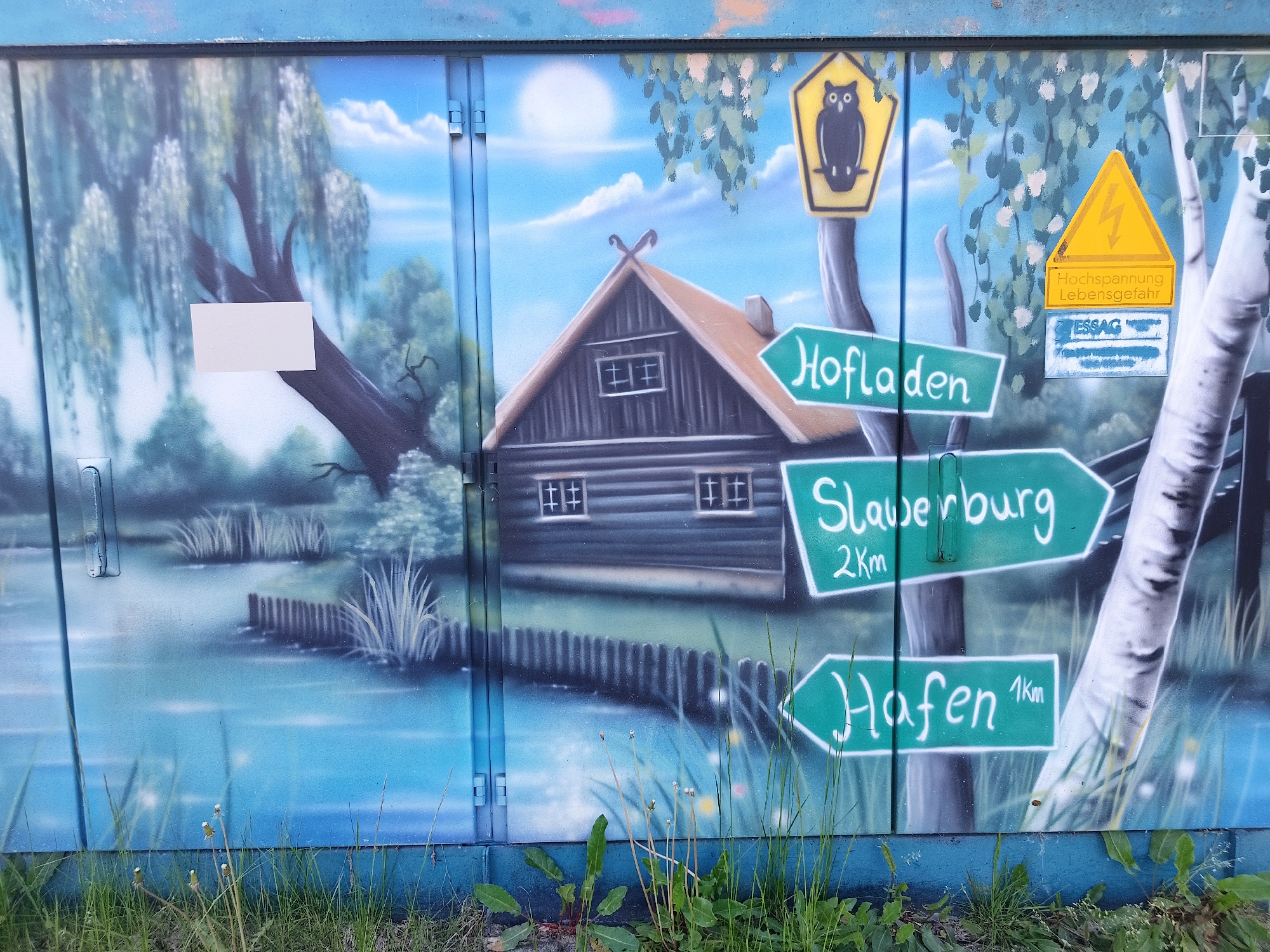 A painted utility box stands amidst overgrown grass in Unknown, Unknown. The painting depicts a serene landscape: a weeping willow tree on the left borders a body of water reflecting a cloudy sky and bright sun. In the center, a small, dark cabin sits near a wooden fence, with a series of green direction signs mounted on a painted tree trunk to the right. The top sign features an owl. The signs point towards "Hofladen," "Slauerburg (2km)," and "Hafen (1km)." Also on the utility box is a yellow triangular warning sign indicating high voltage and life-threatening danger.