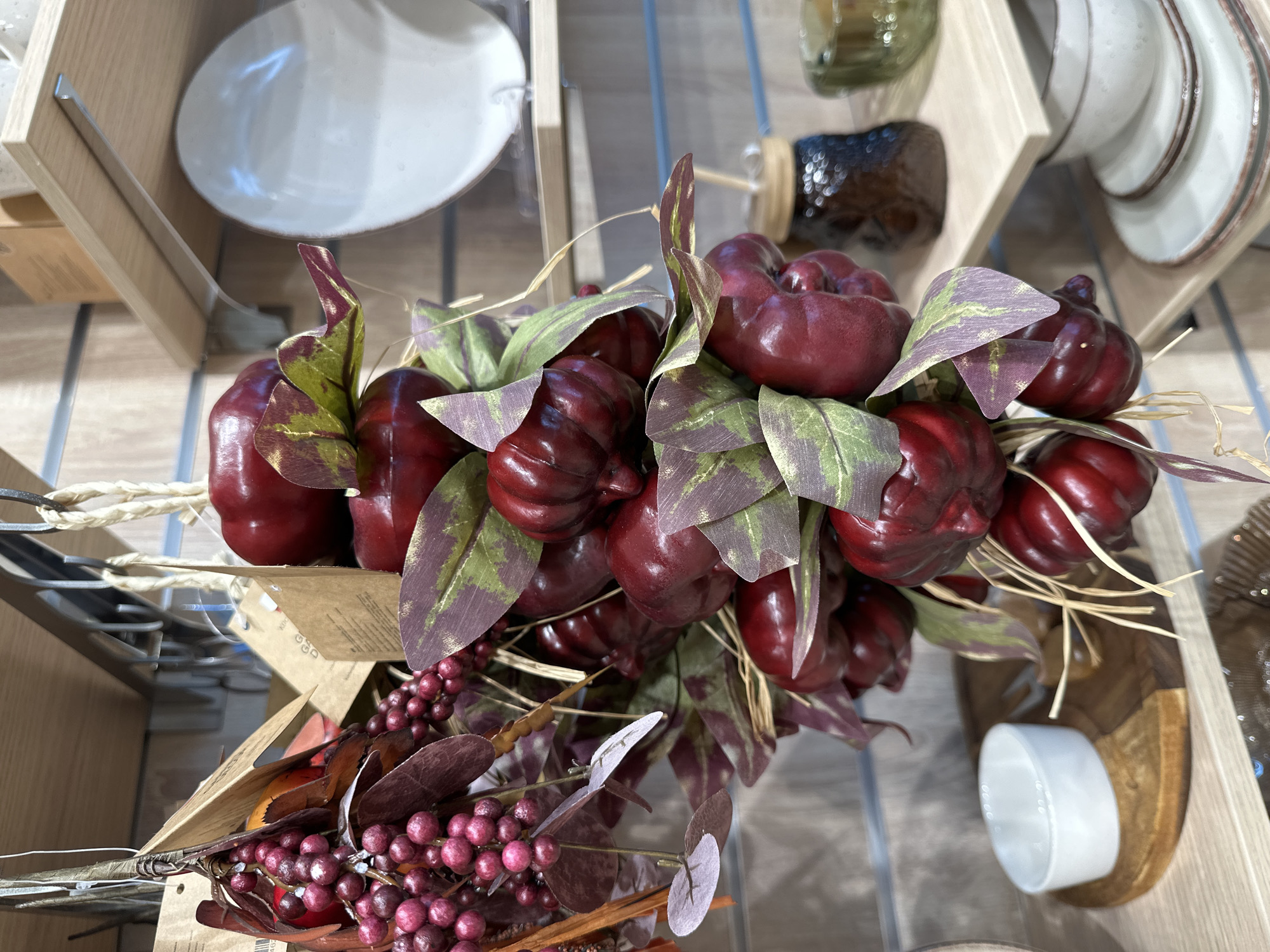 A close-up shot focuses on a decorative arrangement of artificial red fruits and leaves, suspended in front of a display of shelves. The arrangement features numerous small, round, red fruits clustered together, interspersed with dark red and green artificial leaves. Some of the leaves have a distressed or aged appearance, with purple and green hues. A few pieces of straw are also visible, adding to the rustic aesthetic.
Behind the decorative arrangement, wooden shelves are visible. The shelves display various items. On the top shelf, a brown cardboard box is partially visible, and to its right, a white plate or shallow bowl is prominently displayed. The middle shelf shows a dark-colored jar with a wooden lid, and to its right, a green glass object, possibly a candle holder. The bottom shelf contains a stack of white plates with thin brown rims, and above them, a white bowl sits on a two-toned wooden circular base.
The overall environment suggests a retail setting, possibly a home decor or gift shop. The background appears to be a wall with horizontal grooves. There are no people visible in the image, and no actions are being performed. The lighting is even, highlighting the details of the objects. The scene is static and posed.