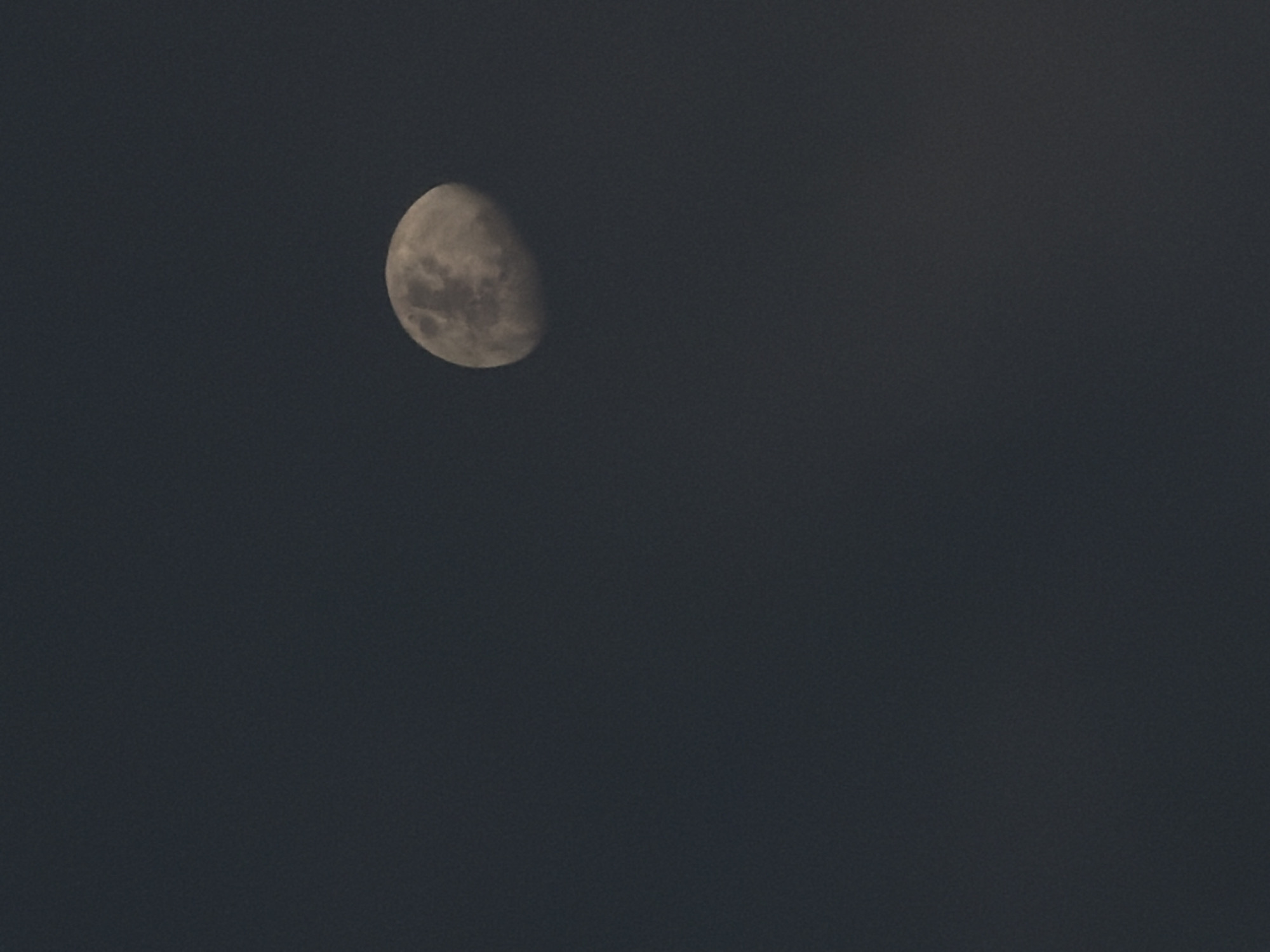 The image shows a half moon in a dark gray sky. There is no evidence to indicate the location is North Kuta, Indonesia. No people, subjects, objects, actions, or interactions besides the moon in the sky are visible. The environment is a dark, seemingly night sky.