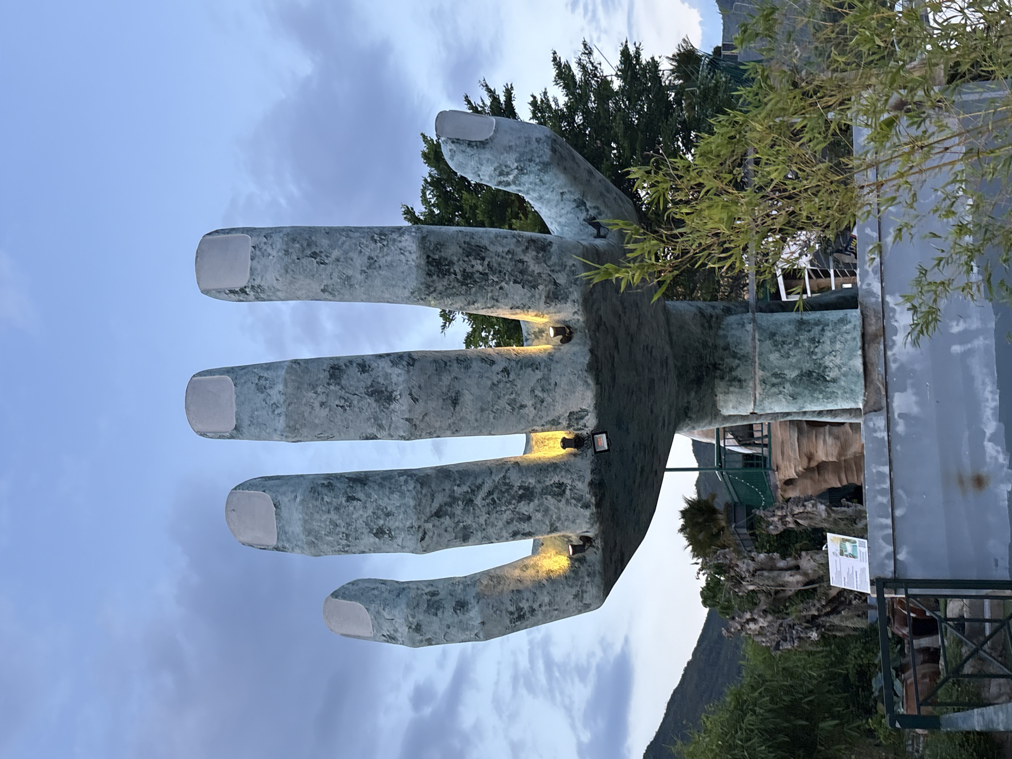 The image shows a large, blue-green, textured sculpture of a hand with five fingers, reaching upwards against a cloudy twilight sky. The sculpture has a rough, somewhat mottled surface, and each fingertip is a lighter, smoother shade, resembling a fingernail. Small, yellow spotlights are attached to the underside of the sculpture, illuminating the palm area. In the background, there are lush green trees, a hillside, and hints of a building structure with railings and an outdoor seating area. A small, white information sign with text and an image is visible on a metal railing in front of some weathered rock formations and large pottery vessels. The overall scene suggests an outdoor art installation or a themed attraction, possibly in a park or garden setting. The location is identified as Nam Nha Trang Ward, Vietnam.