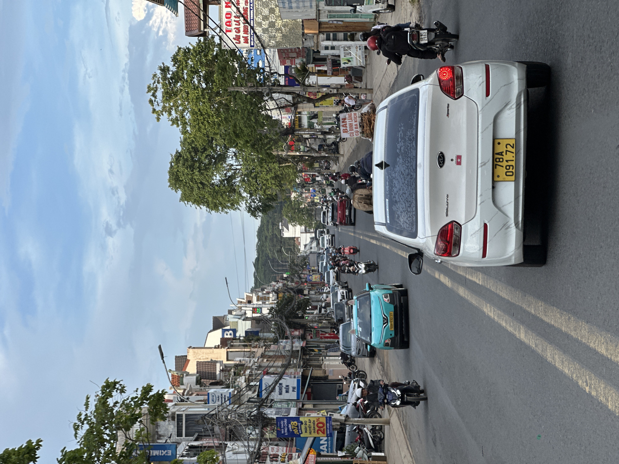 The image depicts a street scene in Lâm Viên - Đà Lạt, Vietnam. The perspective is from within a vehicle, looking forward. The street is paved and appears to be a main thoroughfare lined with two- and three-story buildings on either side, many displaying signs in Vietnamese. The architecture suggests a dense urban environment. Traffic is moderate, consisting of cars, motorcycles, and a few scooters. A white sedan is prominently in the foreground, its rear facing the camera. A turquoise car is visible in the middle distance, as is a line of motorcycles and other vehicles progressing down the road. Trees with green foliage are interspersed along the street, and power lines crisscross overhead. The sky is partially cloudy, with a mix of blue sky and diffused sunlight. The overall impression is of a busy, everyday street in a Vietnamese city.