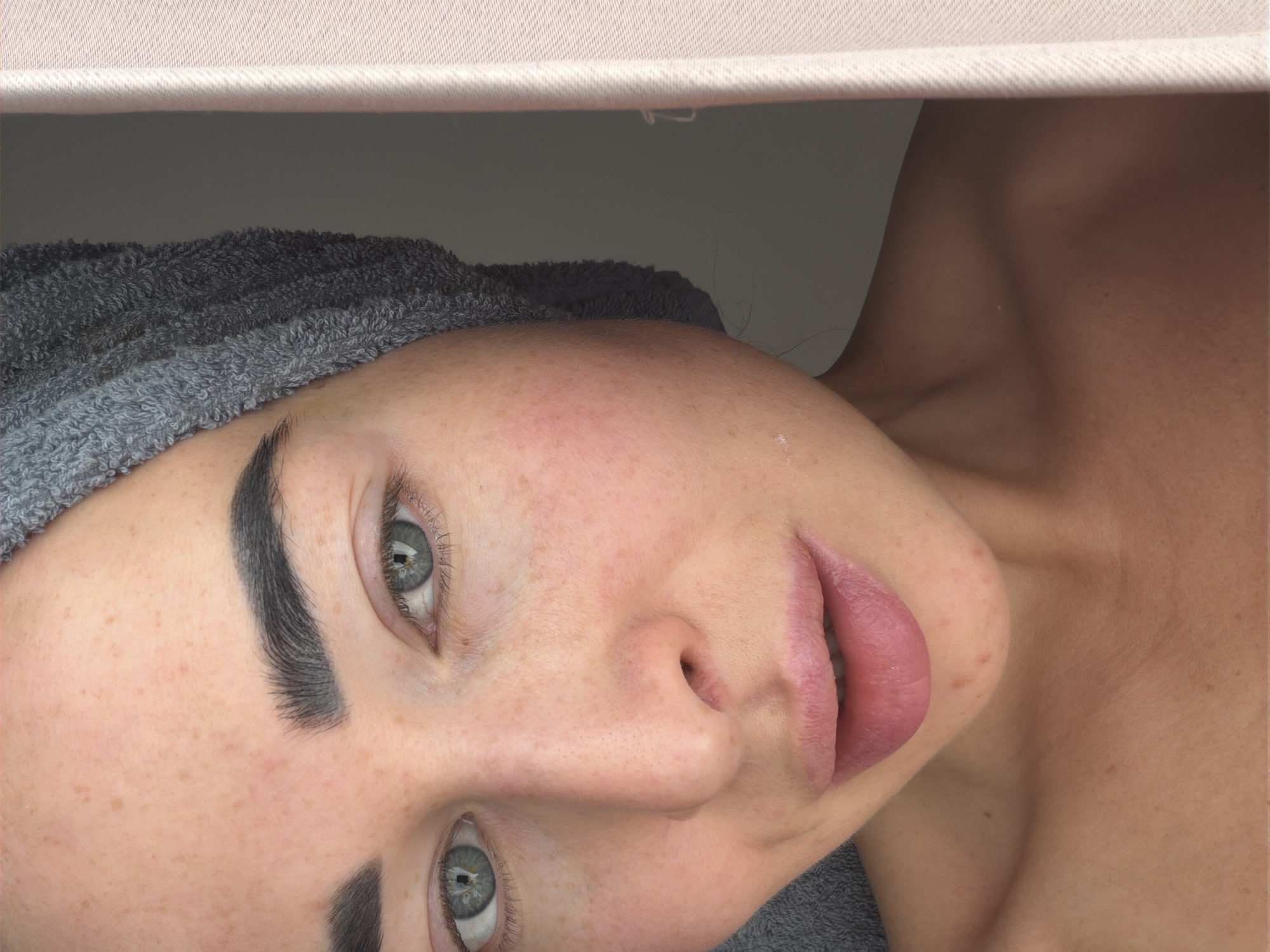 A close-up, horizontally oriented portrait features a person, likely female, with their head resting against a dark grey surface, possibly a wall or headboard. The upper portion of the frame is bordered by a light-colored, textured fabric, resembling a blanket or sheet. The person's hair is wrapped in a soft, grey terry towel.
The subject's face is prominent, characterized by fair skin dotted with numerous subtle freckles across the cheeks, nose, and forehead. Her eyes are a striking blue-grey, open and looking directly towards the camera, with distinct reflections of a light source in her pupils. Her eyebrows are notably thick, dark, and well-defined, providing a strong contrast against her lighter skin. The nose is small and straight, and her full lips, a natural pink shade, are slightly parted. The visible skin on her neck and shoulder also displays freckles and a smooth texture. The lighting is soft and even, casting minimal shadows and creating a serene, calm mood. There is no visible text in the image.