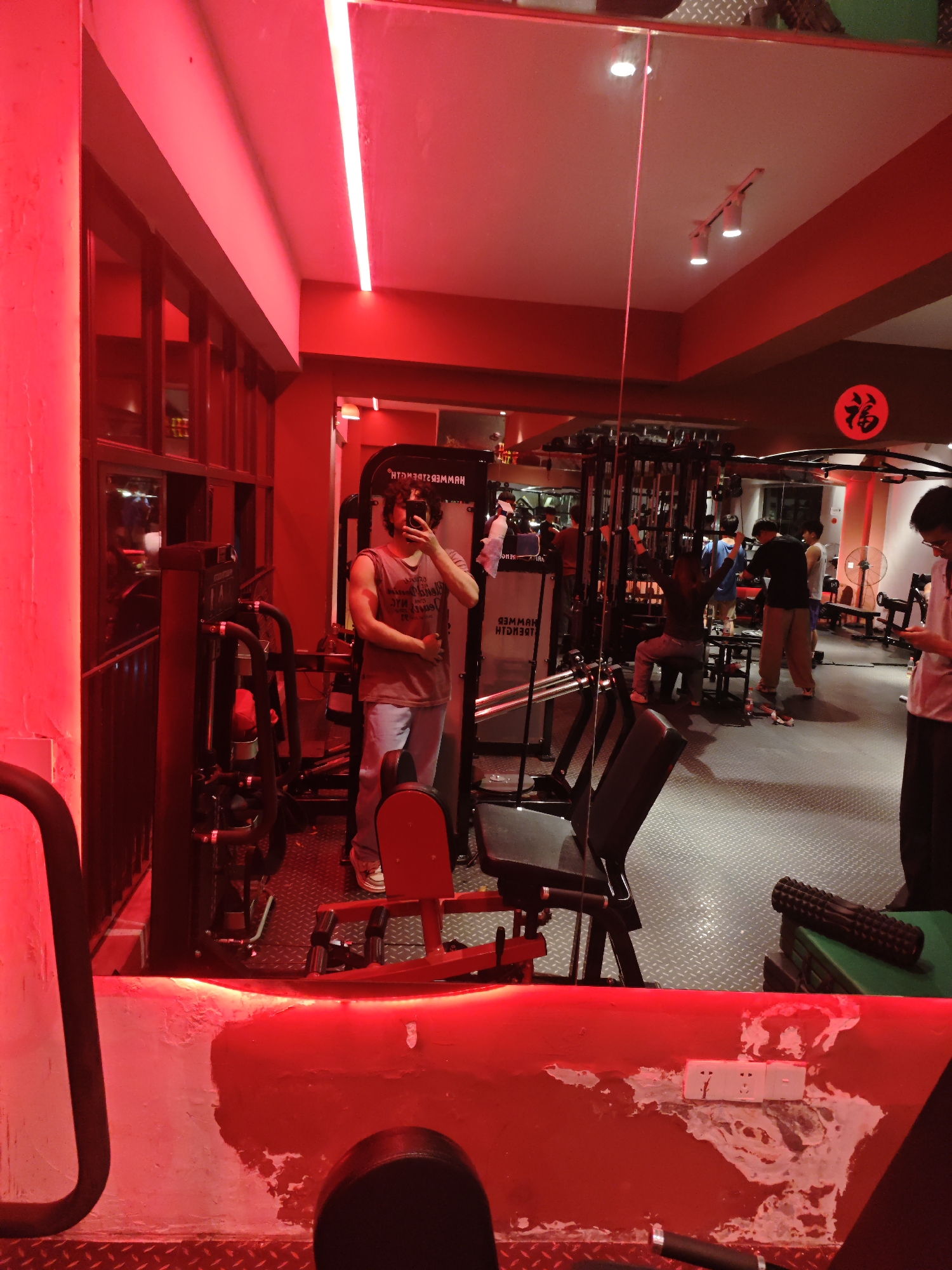 The image shows a person taking a mirror selfie in a gym. The gym has red lighting and various exercise machines, including weight machines, a leg press, and exercise benches. Other individuals are working out in the background. The gym appears to be located in the Chongqing High-tech Zone, China.