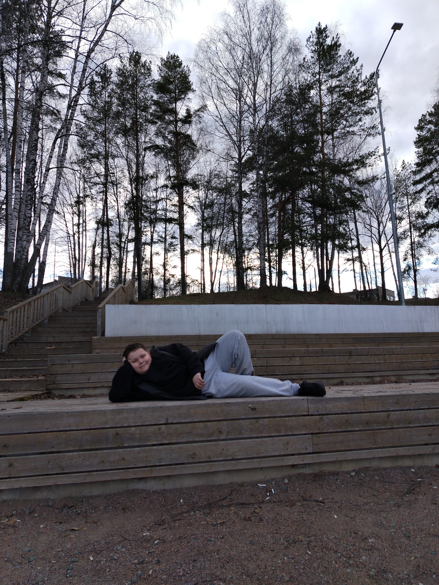 A medium-shot of a young person lying on their side on a wooden structure resembling steps. The individual is wearing a black long-sleeved shirt, grey sweatpants, and black shoes. Their head is resting on their hand, and they appear to be smiling slightly.
Behind the wooden structure is a retaining wall. Above the wall, there is a line of tall trees with sparse foliage, silhouetted against a cloudy sky. To the left of the image are wooden stairs with railings. The ground in the foreground is gravel. A lamppost is visible on the right side of the image.