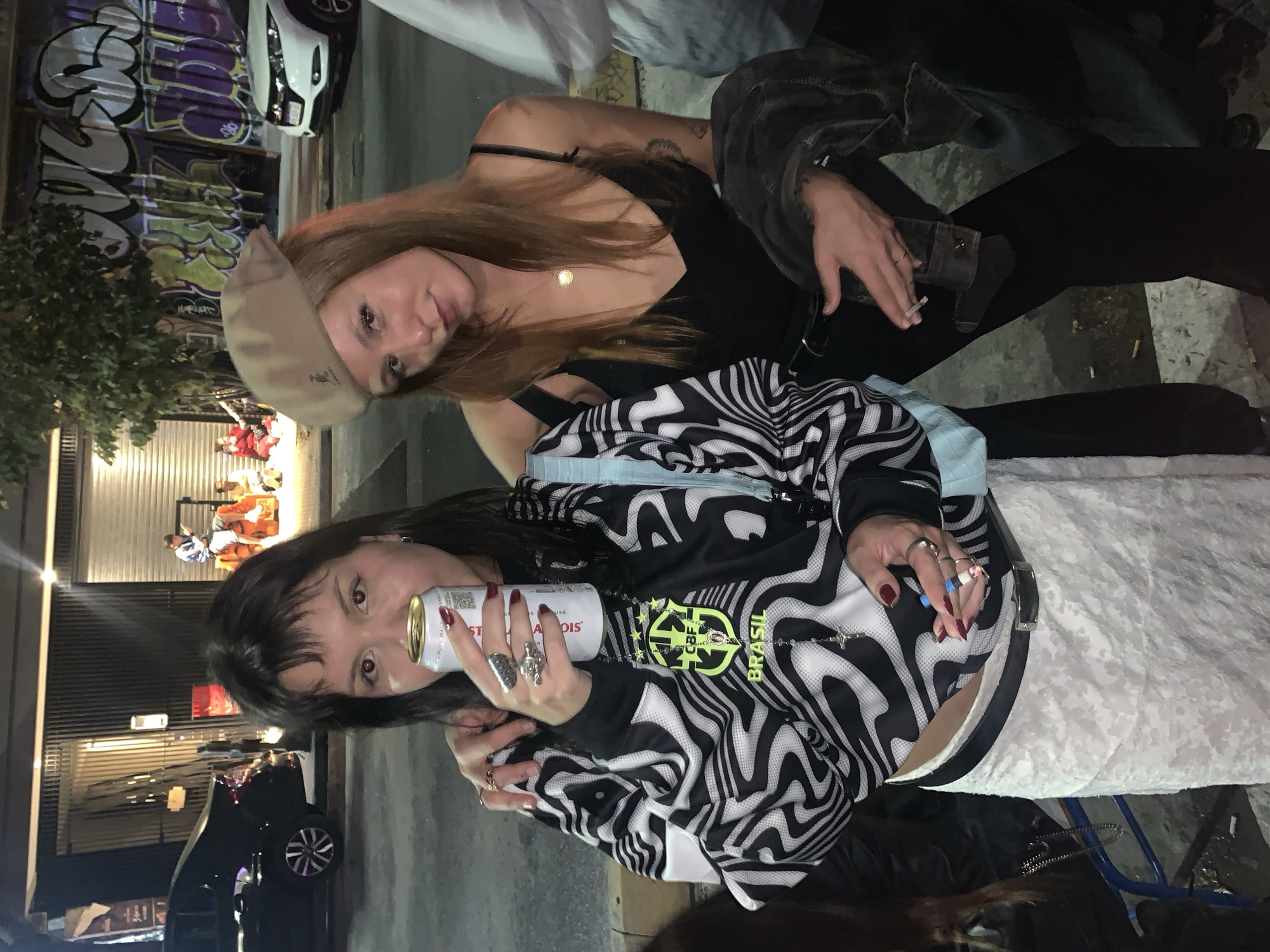 Two young women are posing for a photo outdoors at night, likely in Buenos Aires, Argentina. The woman in the foreground has dark hair and bangs, wearing a black and white zebra-patterned jacket with a light blue shoulder bag. She is holding a can of Stella Artois beer. She has several rings on her fingers, and her nails are painted dark red. She is seated on what appears to be a cushion or a soft surface. The other woman behind her has long reddish-brown hair and wears a beige beret and a black tank top. She has a jacket draped over her shoulders and is holding a cigarette in her hand. In the background, there's a graffitied wall, a parked white car, and a black car. There are also people seated near what looks like a storefront or building. The lighting suggests it is nighttime, and the general setting implies an urban environment.