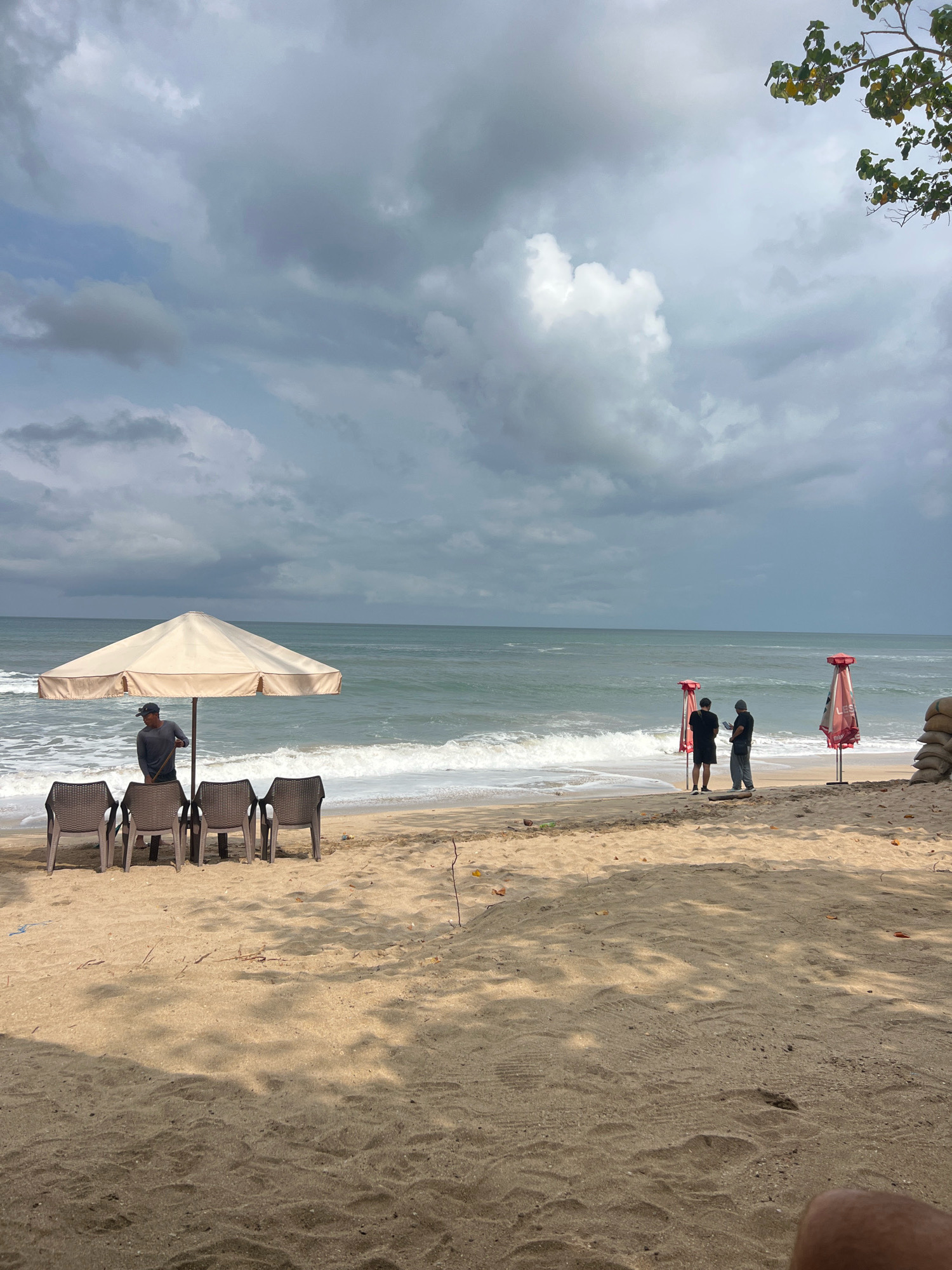 This is a medium shot taken from a sandy beach in Kuta, Indonesia, looking out towards the ocean under a cloudy sky. The foreground is dominated by sand, with some dried leaves and shadows from trees cast across it. In the middle ground, four empty brown plastic chairs are arranged in a row under a large, light-colored beach umbrella. A man wearing a grey long-sleeved shirt and a cap stands behind the umbrella, holding a broom.
Further down the beach, two men in casual attire, one in a black t-shirt and shorts, the other in a grey t-shirt and pants, stand near the water's edge. They appear to be interacting with each other, possibly looking at a phone or a piece of paper. Two closed red and white beach umbrellas are planted in the sand behind them. In the far background, the ocean is visible with moderate waves breaking on the shore, and the horizon line is indistinct due to the overcast sky. To the right, a small stack of sandbags is visible. The overall atmosphere suggests a quiet, possibly pre-storm, day at the beach.