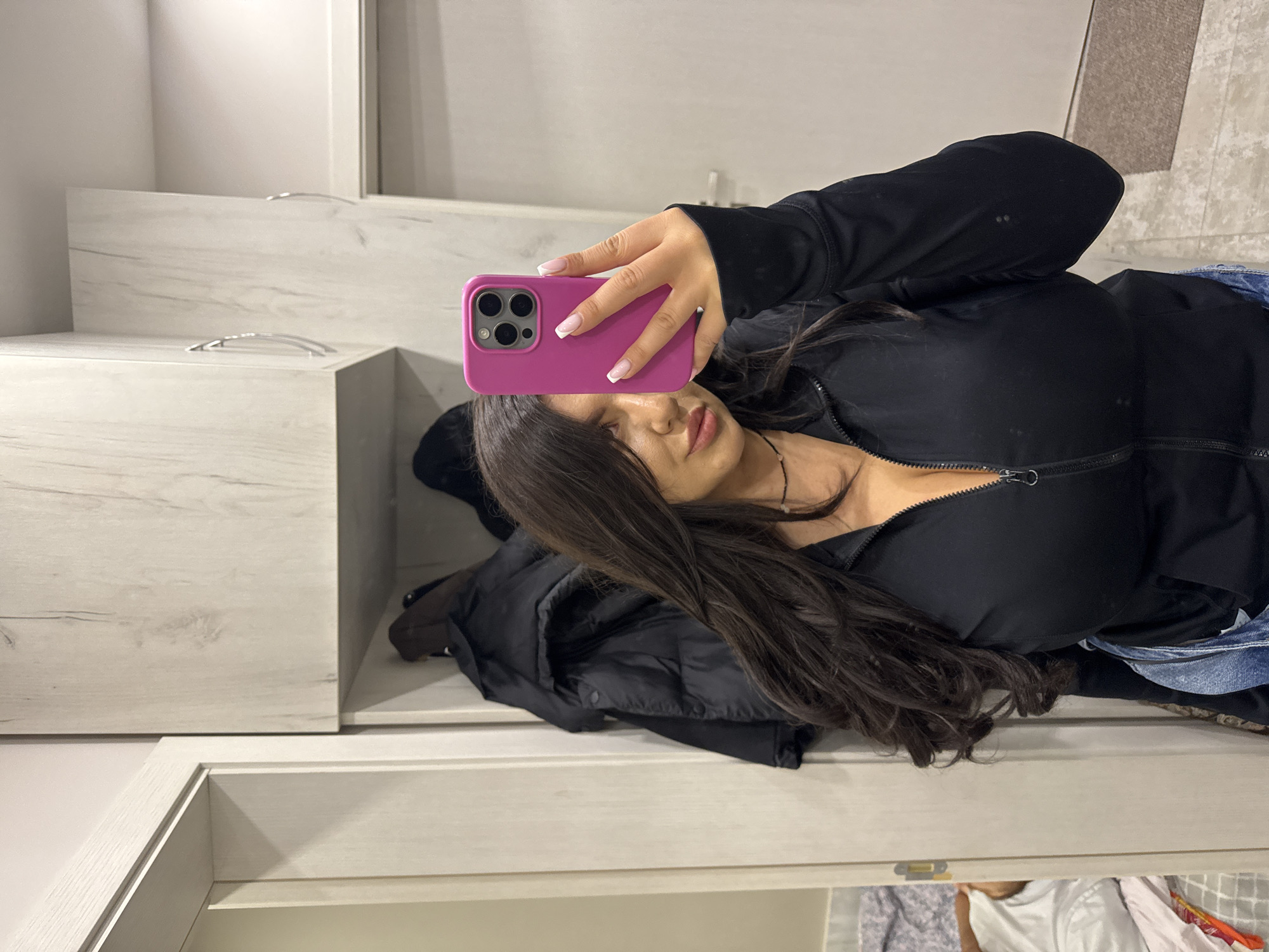 A medium-shot selfie shows a woman indoors, likely in a residential setting. She is holding a pink smartphone in her right hand, taking a photo in front of a mirror. The woman has long, dark brown hair and is wearing a black zippered jacket. Her manicured nails are visible. Behind her, there is a wardrobe or closet with light-colored, wood-grain textured doors. A black jacket or coat is draped over the edge of an open drawer. Below the open drawer, clothing is visible in a lower drawer or compartment. The location is not identifiable as Burgas, Bulgaria based on the image.