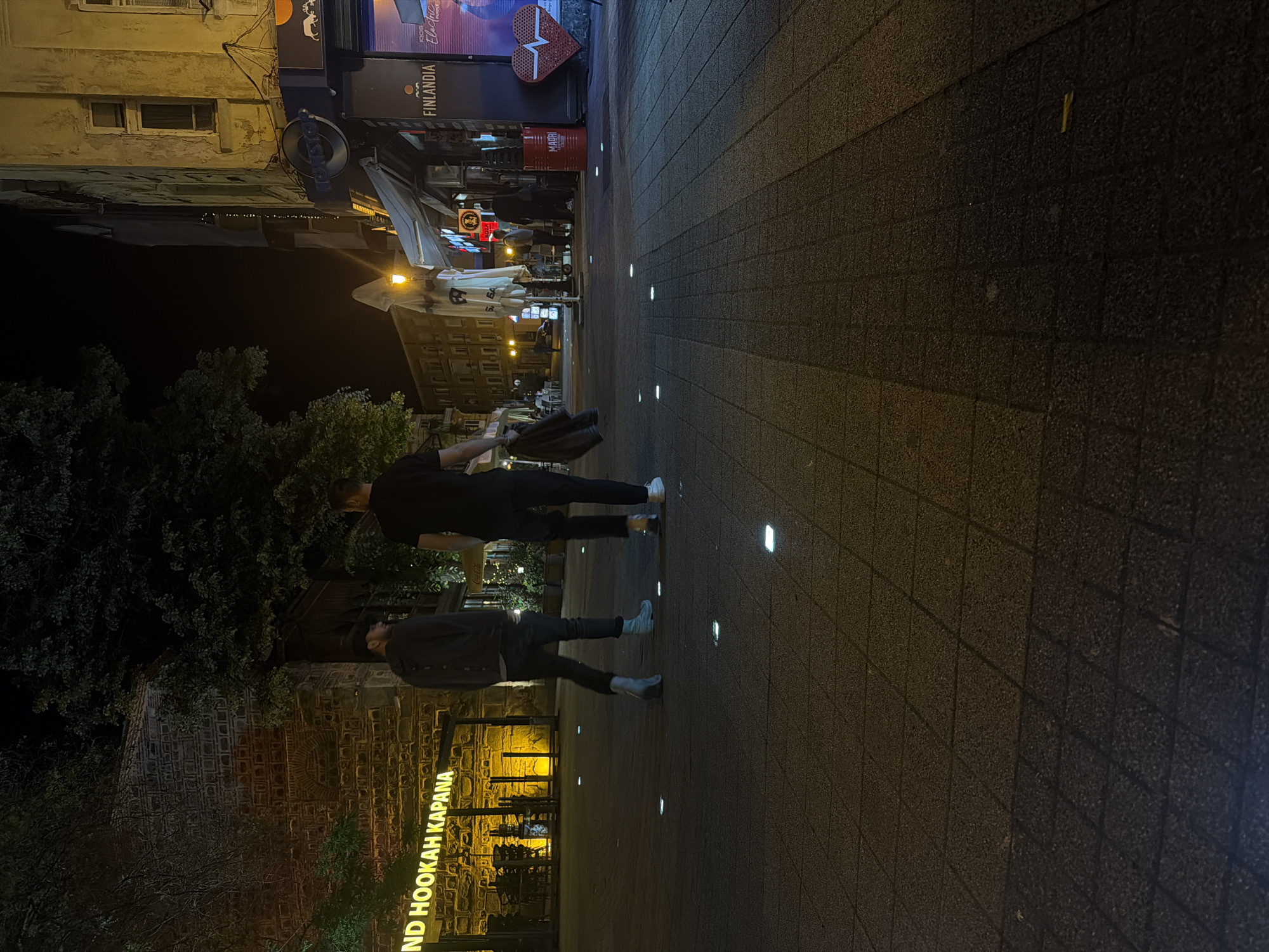 Here's a description of the image:
**Location:** The image depicts a nighttime street scene in Plovdiv, Bulgaria, specifically in the Kapana district. "KAPANA" is visible on a sign on the bottom left corner of the frame.
**People:** There are at least two people present. One appears to be carrying a bag. Both people are walking.
**Objects:** The scene includes buildings with illuminated signage (Finladia), trees, street lighting embedded in the paving, and a paved street or square.
**Environment:** It is nighttime, and the scene is artificially lit. The paving is made of dark, square tiles.