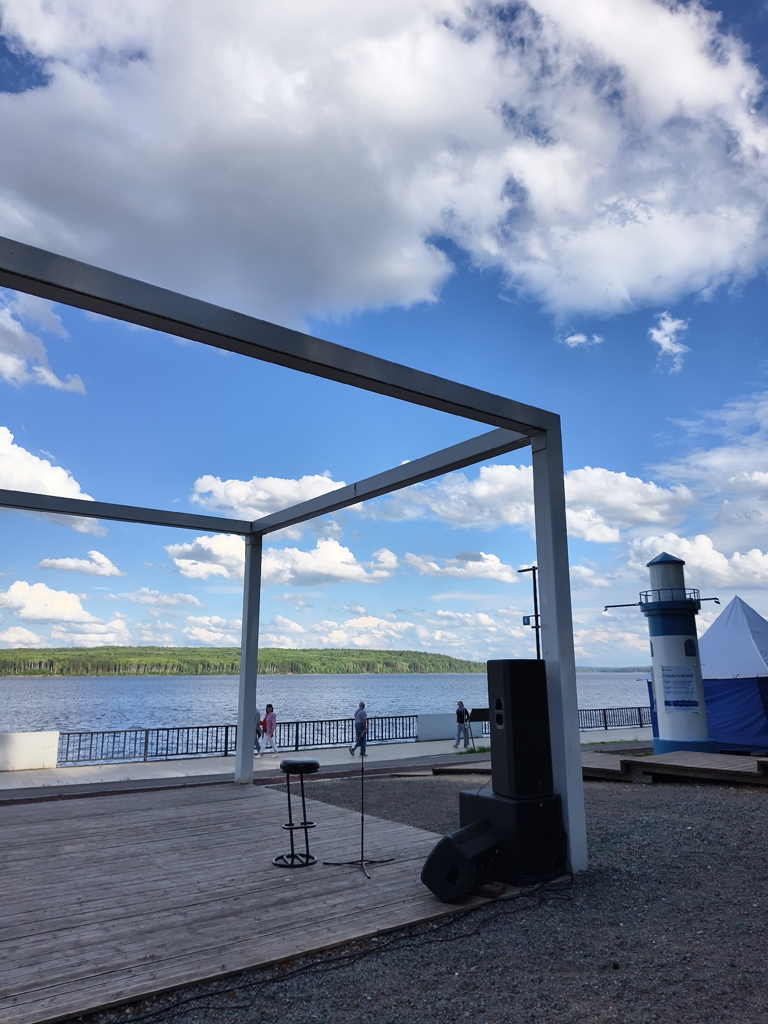 A wide shot captures a view of a calm lake on a partly cloudy day. In the foreground, a wooden stage is set up, with a bar stool and two large black speakers positioned on it. A metal frame structure extends from the right side of the image, partially obscuring the sky. Beyond the stage, a paved walkway runs alongside the lake, bordered by a black metal railing. Several people are walking along the walkway. In the distance, a white and blue lighthouse stands tall, and a white tent is visible to its right. The far bank of the lake is lined with a dense green forest.