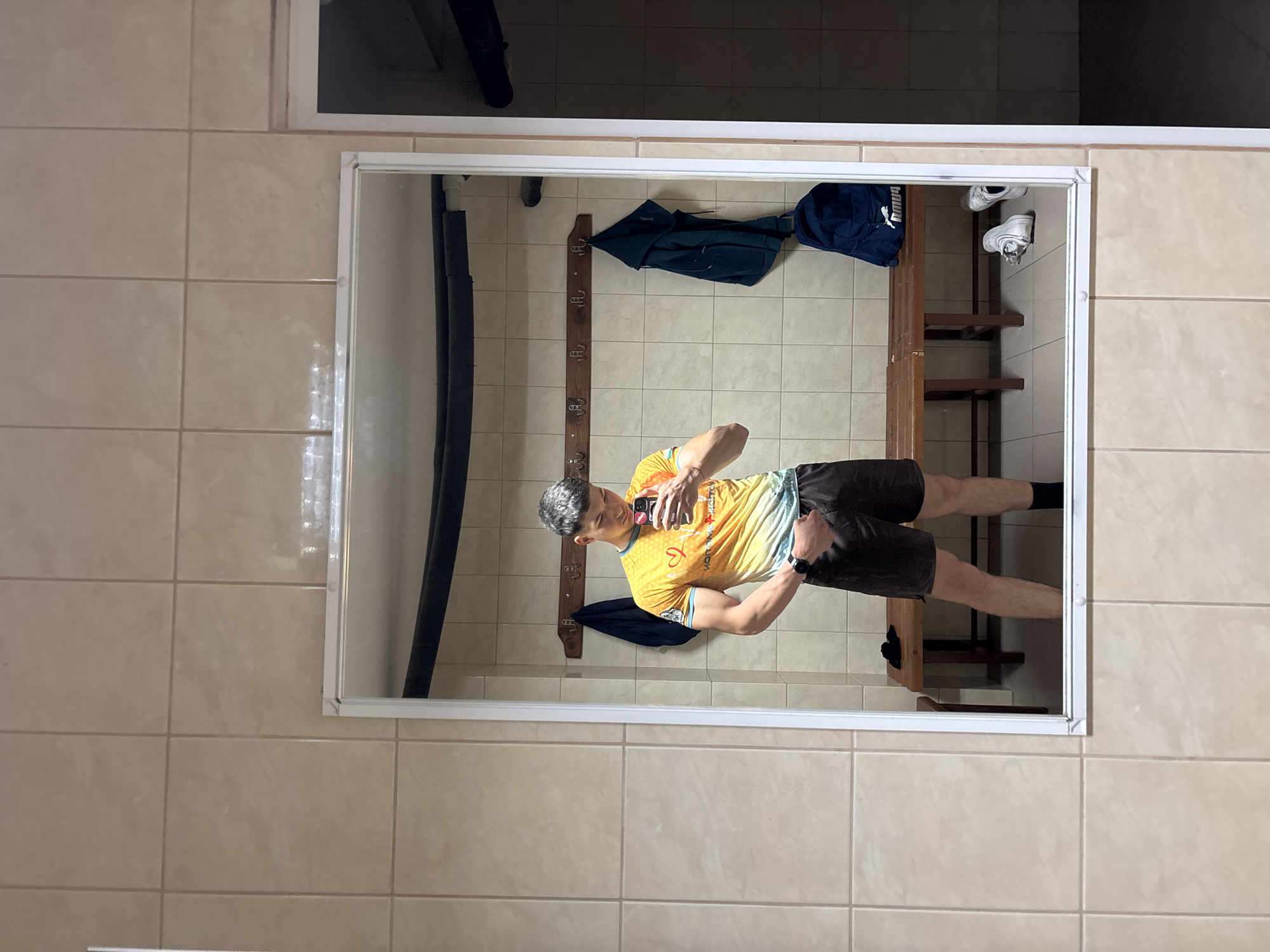 A man is taking a selfie in a mirror in a locker room. The man is wearing a yellow and light blue t-shirt and dark shorts. He is holding a smartphone in his right hand. The locker room has beige tile walls and floor. There is a bench with a blue bag and white sneakers on top of it. There is also a coat rack with clothing hanging on it. The locker room is located in Zlatograd, Bulgaria.