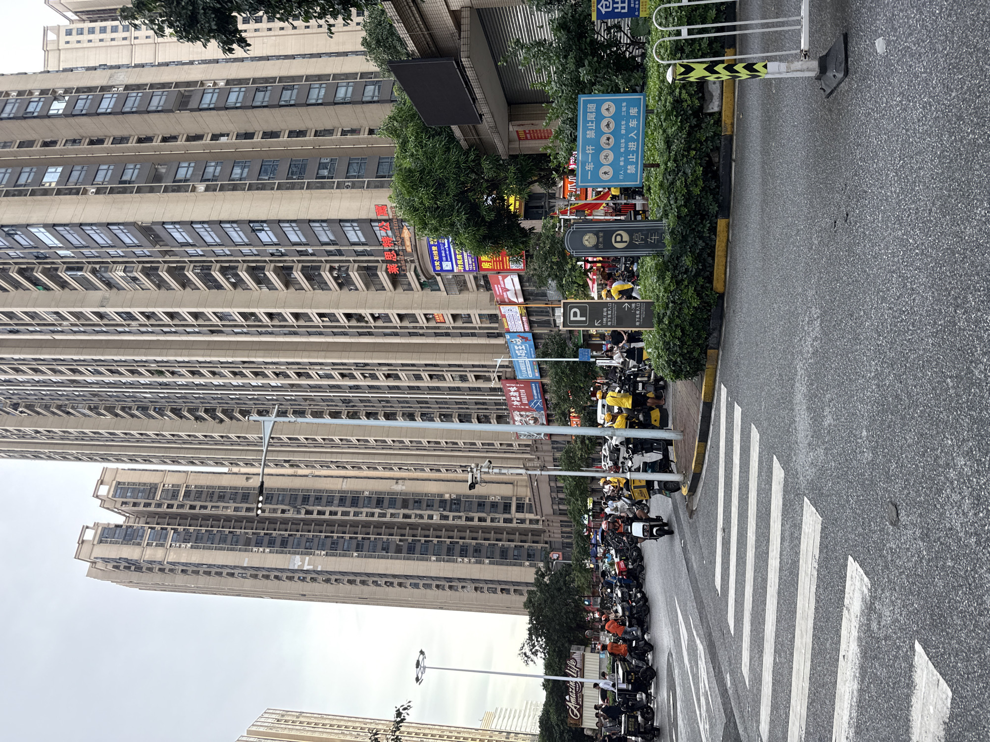 This is a vertical, eye-level medium shot of a street scene in Haizhu District, China. The dominant subjects are towering apartment buildings and a row of parked electric scooters and motorcycles along the side of a street. In the foreground, a crosswalk is visible, marked by white stripes on asphalt.
The scene is set on an overcast day, suggested by the diffused lighting and muted colors. The apartment buildings are multi-story structures with numerous windows, indicative of a densely populated urban environment. Along the sidewalk, several signs are visible, some with Chinese characters and parking symbols. One sign in particular states "禁止进入车库" (Forbidden to enter garage) and also lists prohibited vehicles like pedestrians, bicycles, electric scooters, motorcycles, and tricycles. Another sign advertises "Amazin Life."
The parked scooters and motorcycles suggest that this is a common mode of transportation in the area. Some of the scooters are yellow, likely delivery vehicles. People are present, but their faces are not clearly discernible, suggesting they are either moving or their features are obscured. The overall atmosphere is one of urban activity and routine. The presence of multiple signs and the density of buildings point towards a commercial or residential district.