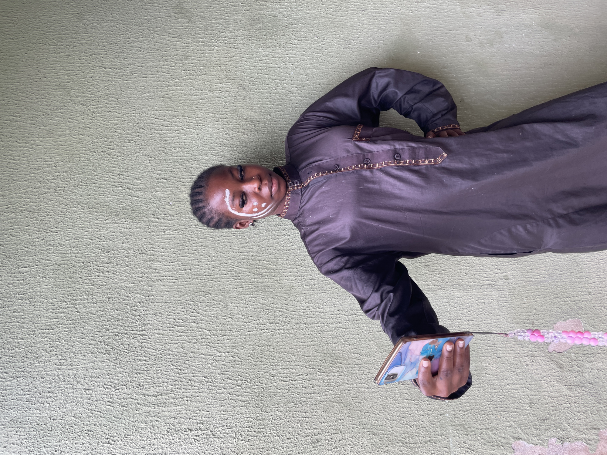 A young Black individual with braided hair and white face paint is standing against a light green textured wall, holding a smartphone in their right hand. The individual is wearing a dark, long-sleeved garment with gold trim. The smartphone has a colorful case and a pink beaded charm attached. The wall appears to be an exterior surface, possibly of a building. The lighting suggests it is daytime. The image is taken from a medium shot, focusing on the individual and their immediate surroundings. The specific location within Ado, Nigeria, cannot be determined from the visual information provided.