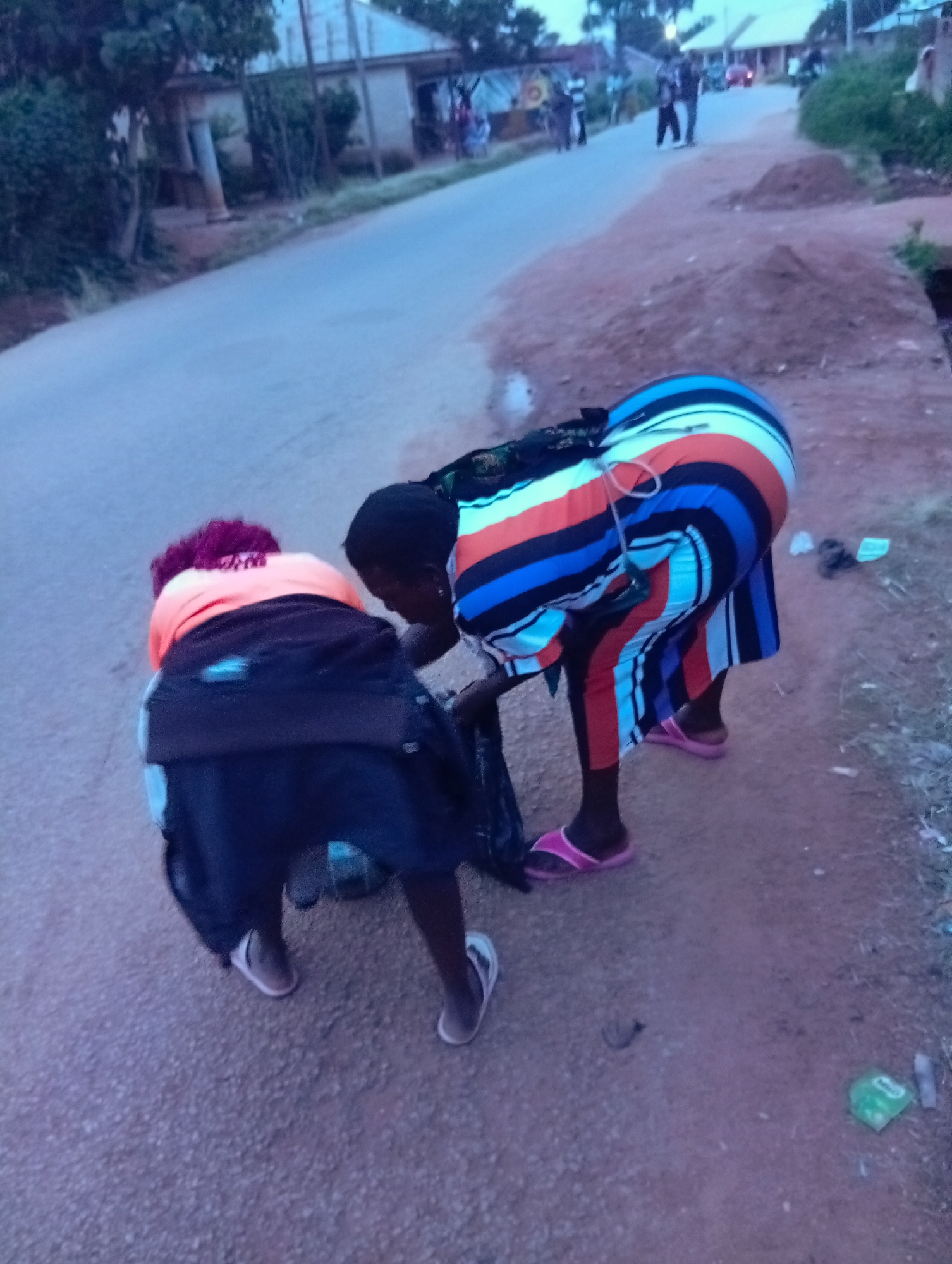 Two individuals are bending down near a road in Du, Nigeria. The person on the left has reddish-pink hair and is wearing an orange top and a dark jacket. The person on the right is wearing a multicolored striped dress and pink sandals. They appear to be interacting with something on the ground, possibly collecting trash. The environment suggests a rural or semi-urban setting with a dirt shoulder bordering a paved road. Buildings and trees are visible in the background, and the lighting indicates it is either early morning or late evening.