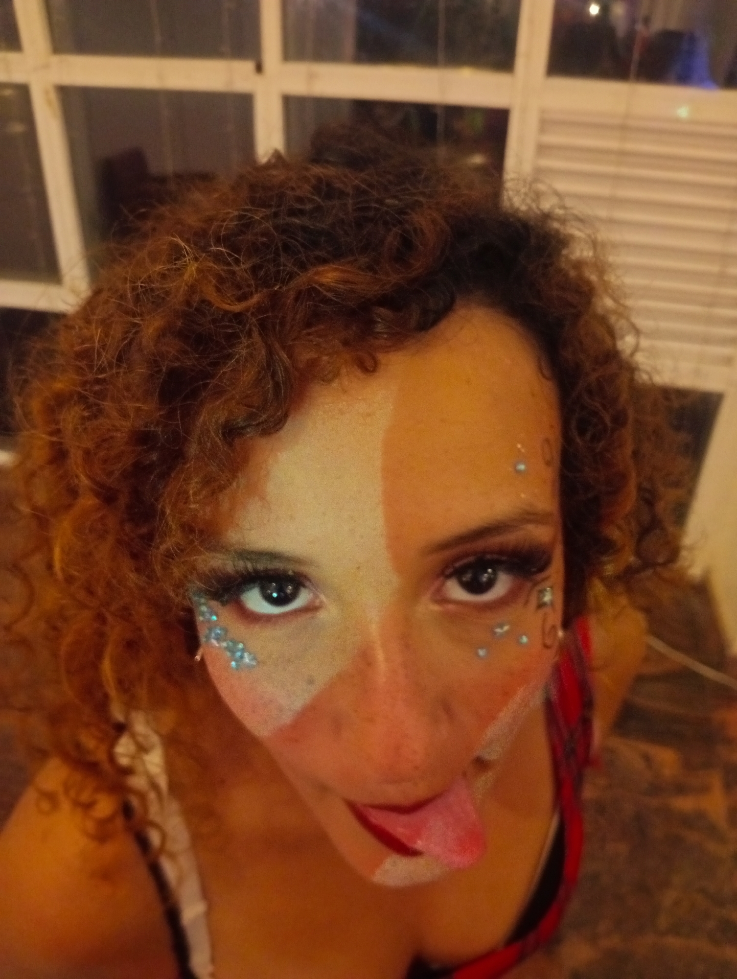 A close-up, high-angle photograph depicts a woman with curly red-brown hair, fair skin, and a split-face makeup design. The left side of her face is bare except for freckles and small, reflective, light-blue adornments clustered around the eye. The right side of her face is covered in a lighter-colored makeup extending from the forehead to around the nose, with similar light-blue adornments. The woman's tongue is extended, coated with lipstick. She appears to be wearing a top with a red plaid pattern.
The background features a multi-pane window, suggesting an indoor location. The image's location within the city of Pedro González, Venezuela cannot be determined from the information provided.
