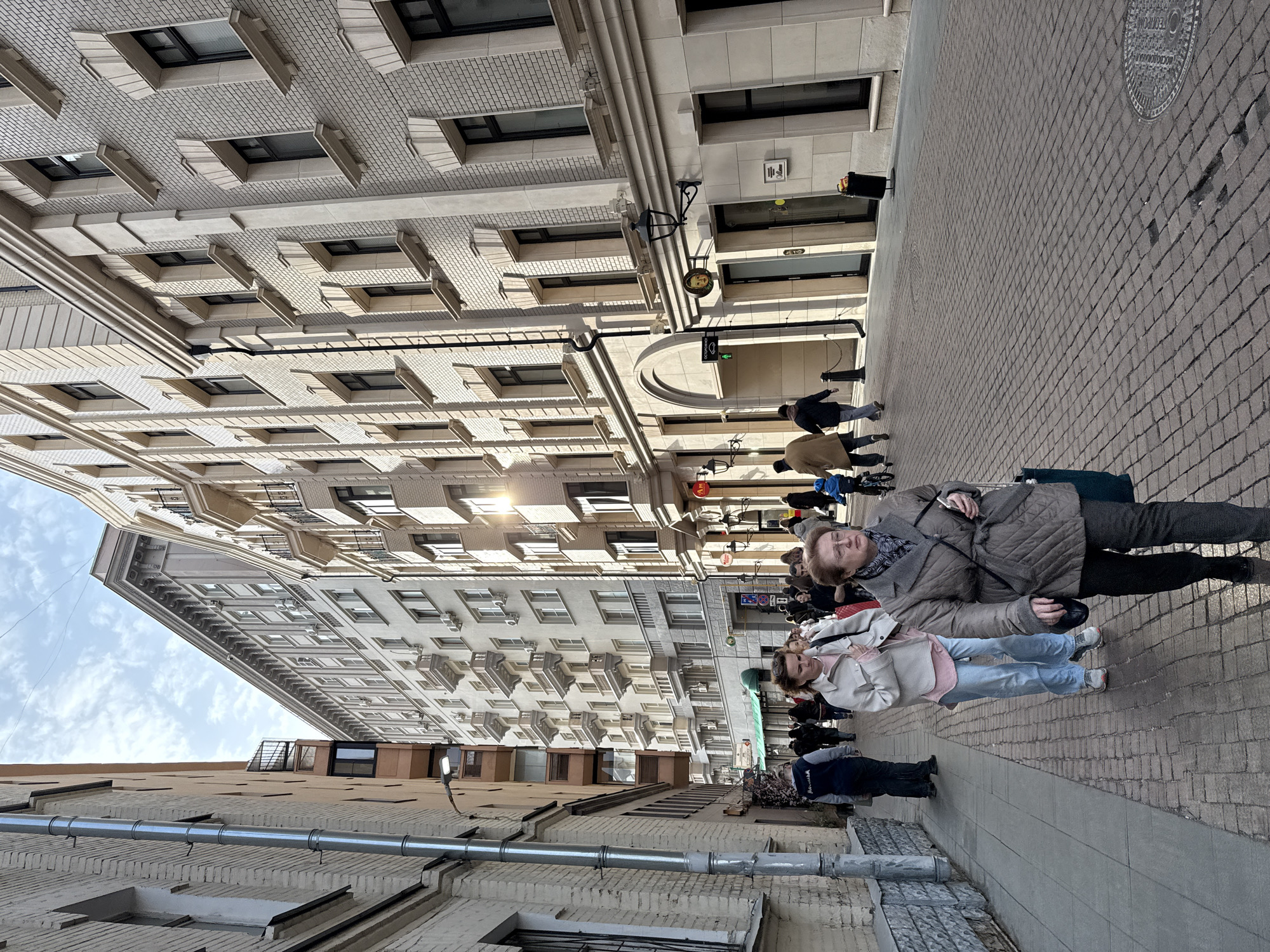 This image depicts a pedestrianized street identified as Bolshaya Dmitrovka Street in Moscow, Russia. The scene is observed under a partly cloudy sky with direct sunlight, causing significant glare on the facades of the buildings and casting shadows.
The street is paved with dark grey cobblestones. On the left, an older, multi-story building with light-colored brick exhibits a vertical metal downspout and multiple windows. On the right, a multi-story building features cream-colored, textured brickwork, numerous windows with decorative light-colored surrounds, an arched entrance on the ground floor, and several retail storefronts, one marked by a circular yellow sign depicting a stylized face and another displaying the Cyrillic letters "Am". In the background, a taller, multi-story building is visible, characterized by numerous small balconies and external air conditioning units.
A moderate number of people are present on the street, walking in various directions. In the foreground, an older woman with glasses, wearing a brown quilted jacket, dark trousers, and a patterned scarf, carries a dark shoulder bag and a mobile phone while walking towards the viewer on the right side of the street. Behind her, a younger woman in a light grey jacket, pink shirt, and light blue jeans walks in the same direction, carrying a white bag. Other individuals, including a person pushing a child in a blue stroller/wagon and a person in a dark blue hooded jacket on the left sidewalk, are distributed in the midground and background, all engaged in walking.
A black cylindrical trash bin is positioned on the right sidewalk. A single street lamp extends from the left building. A circular metal manhole cover is visible on the cobblestone street in the lower right.