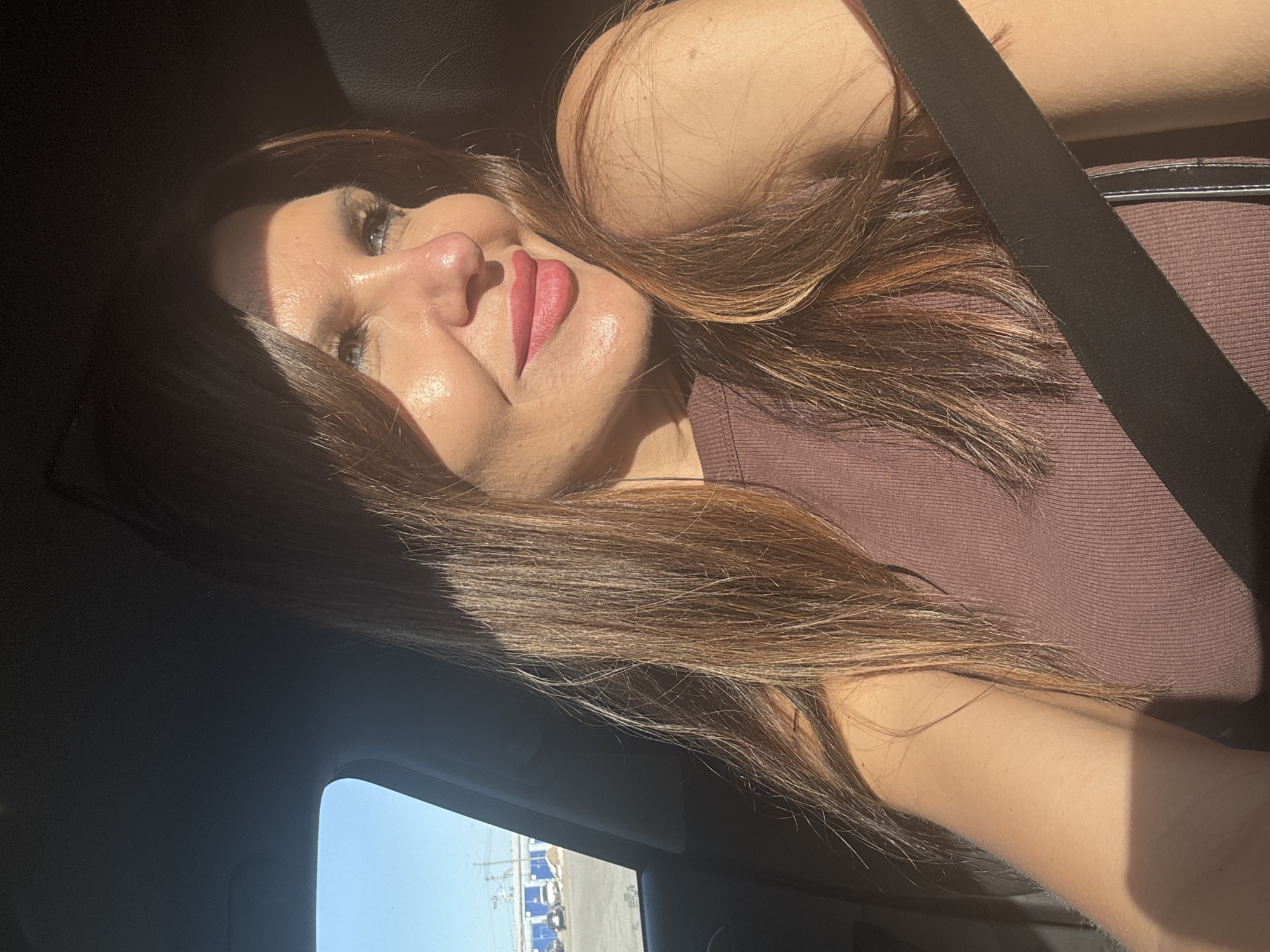 A woman with long brown hair is sitting in a car. She is wearing a brown top and a seatbelt. The sun is shining brightly. Through the car window, there are several cars in a parking lot outside of a building. This photo was not taken in Chaikovsky, Russia.