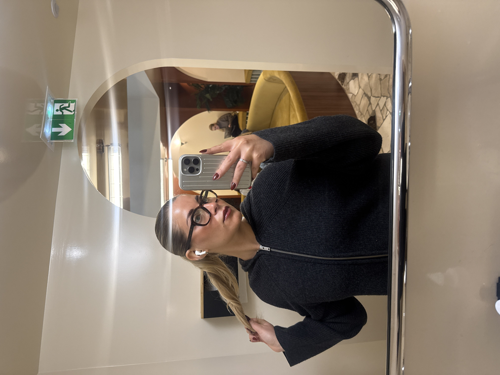 A woman is taking a selfie in a mirror. The mirror is framed with a curved, stainless-steel frame. The woman is wearing glasses and a black zip-up sweater. She is holding her phone in her right hand, which has dark red nail polish. She has a blonde ponytail, and is wearing a white wireless earbud in her left ear.
The reflection in the mirror shows what appears to be an airport lounge. There is a yellow, egg-shaped chair and a person sitting in the background. An exit sign is visible on the wall to the left of the mirror. Given the decor and likely use of the lounge, the location might be Charles de Gaulle Airport, Paris, France.