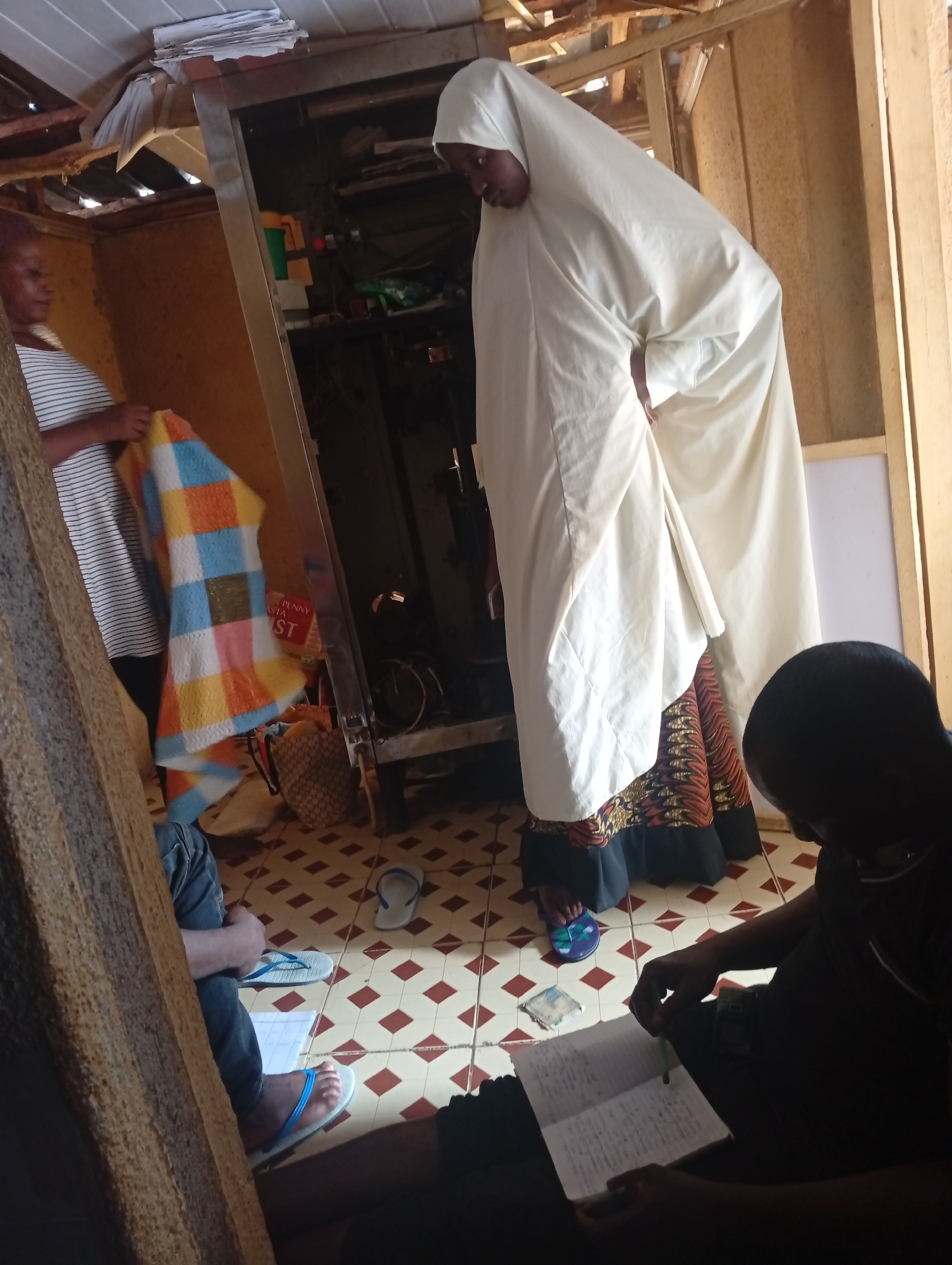 The media file shows an indoor scene with three people. Two women are standing, and one person is seated. The standing women are wearing traditional African clothing, one in a white hijab and the other in a striped shirt. One of the standing women is holding a colorful blanket. The seated person, wearing dark clothing, is looking down at a notebook and a pen, seemingly engaged in writing or reading. The room appears to be modestly furnished, with a tiled floor and unfinished wooden structures visible. The lighting suggests it could be daytime. In the background, there are shelves with various items, including what looks like a basket and some papers. A sign with visible text partially obscured by a bag says "PENNY ASTA ST". The overall impression is of a casual, possibly domestic or educational setting in Jos, Nigeria.