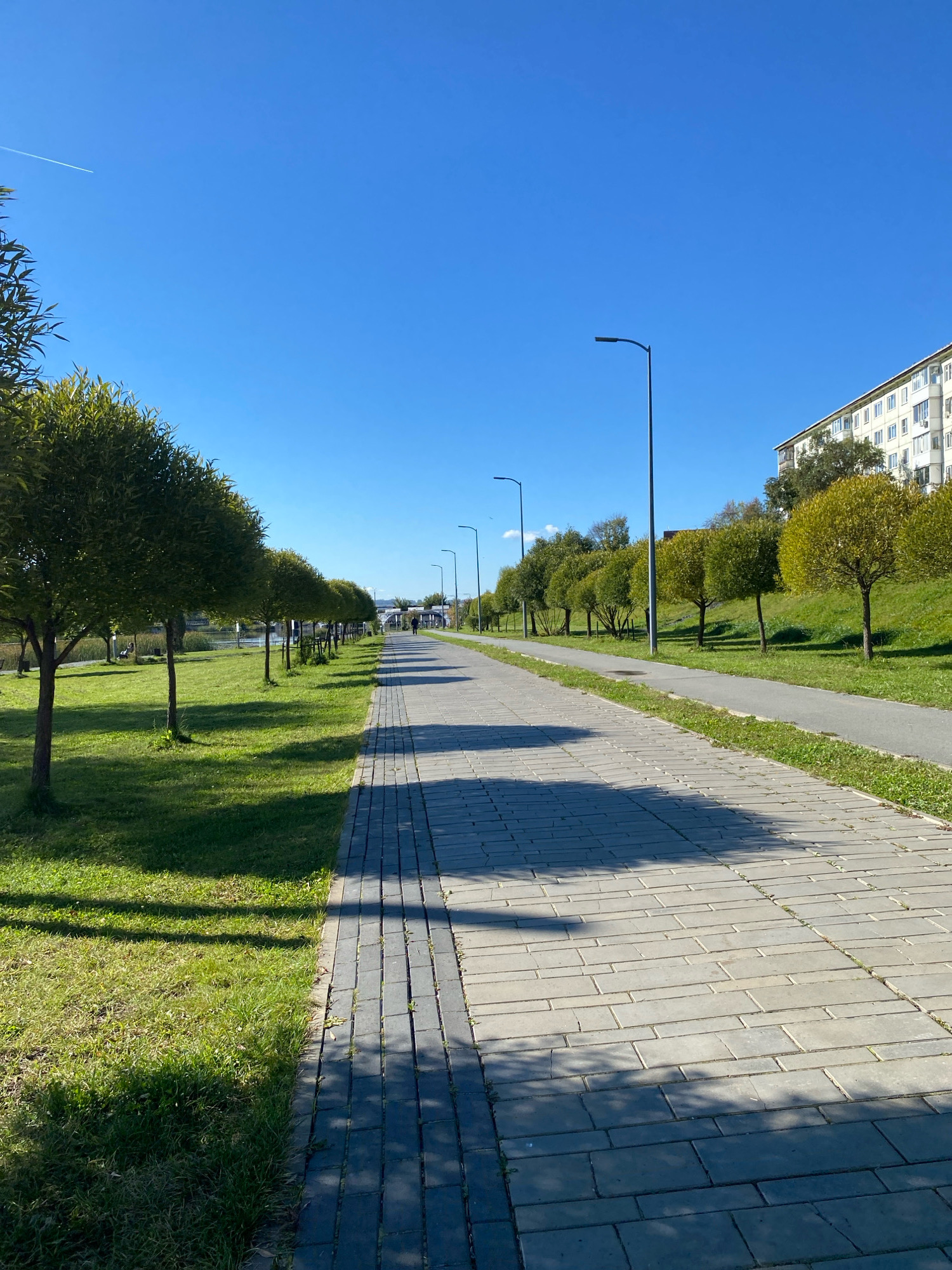 The image depicts a sunny day in Votkinsk, Russia, with a clear blue sky and a row of trees lining a paved walkway. The walkway is made of grey pavers and is bordered by a grassy area on the left, where more trees are planted. To the right of the walkway, there is a slope covered in grass, leading up to a multi-story residential building with many windows. Streetlights are visible along the walkway, spaced at regular intervals. In the distance, a person can be seen walking along the path. A faint white contrail from an airplane is visible in the upper left corner of the sky. The overall scene suggests a well-maintained urban park or promenade.