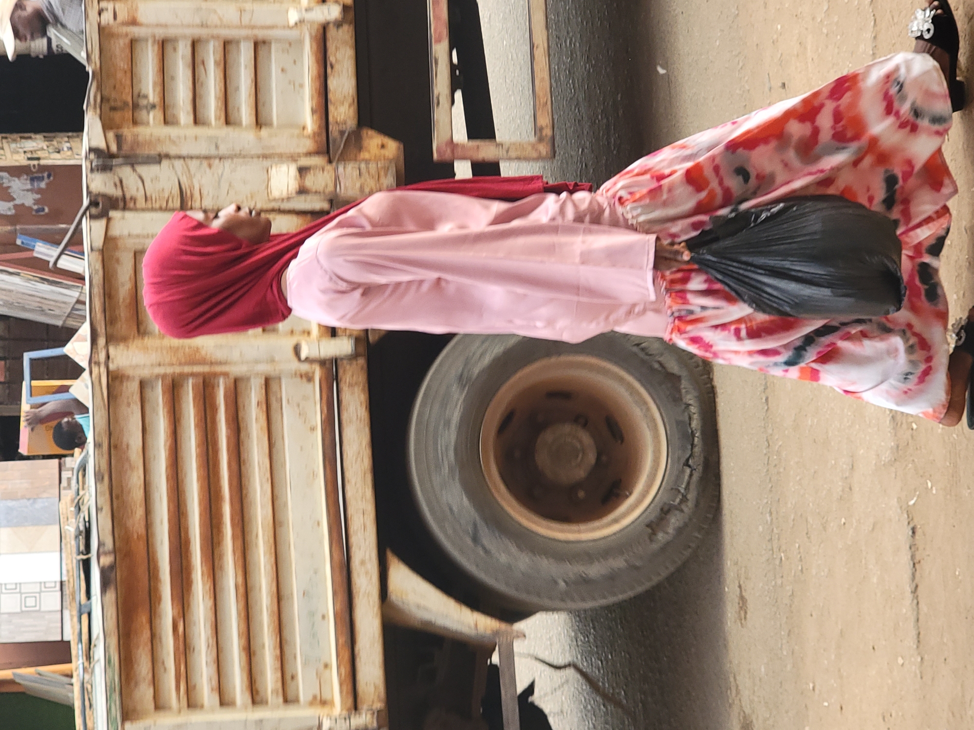 A woman in a red hijab, pink shirt, and tie-dye skirt is standing near a large truck wheel on a street in Maiburiji, Nigeria. She is holding a black plastic bag. The truck has a rusty, white cargo container. There is a building visible in the background.