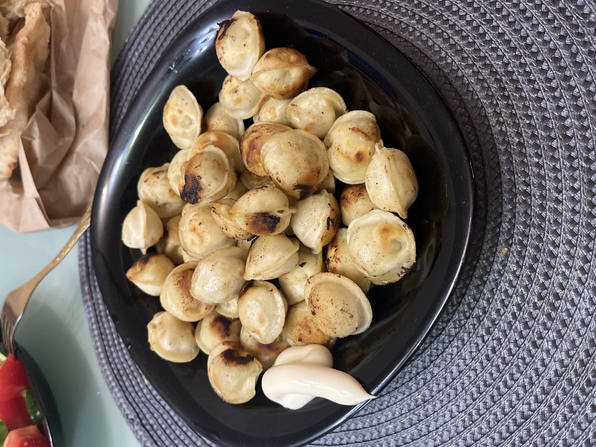 A black plate of fried dumplings, possibly pelmeni, sits on a dark grey woven placemat. The dumplings are golden brown with some darker, almost burnt spots on their surfaces. They are arranged in a pile, and a dollop of white sauce, likely mayonnaise or sour cream, is placed at the bottom right of the plate. To the left of the plate, partially visible, is a crumpled brown paper bag, suggesting a casual dining setting. A silver fork is also visible on the left edge of the image, with a small portion of a salad containing tomatoes and cucumbers. The overall scene appears to be a close-up shot of a meal, likely taken indoors. The lighting suggests it is daytime. There is no visible text in the image.