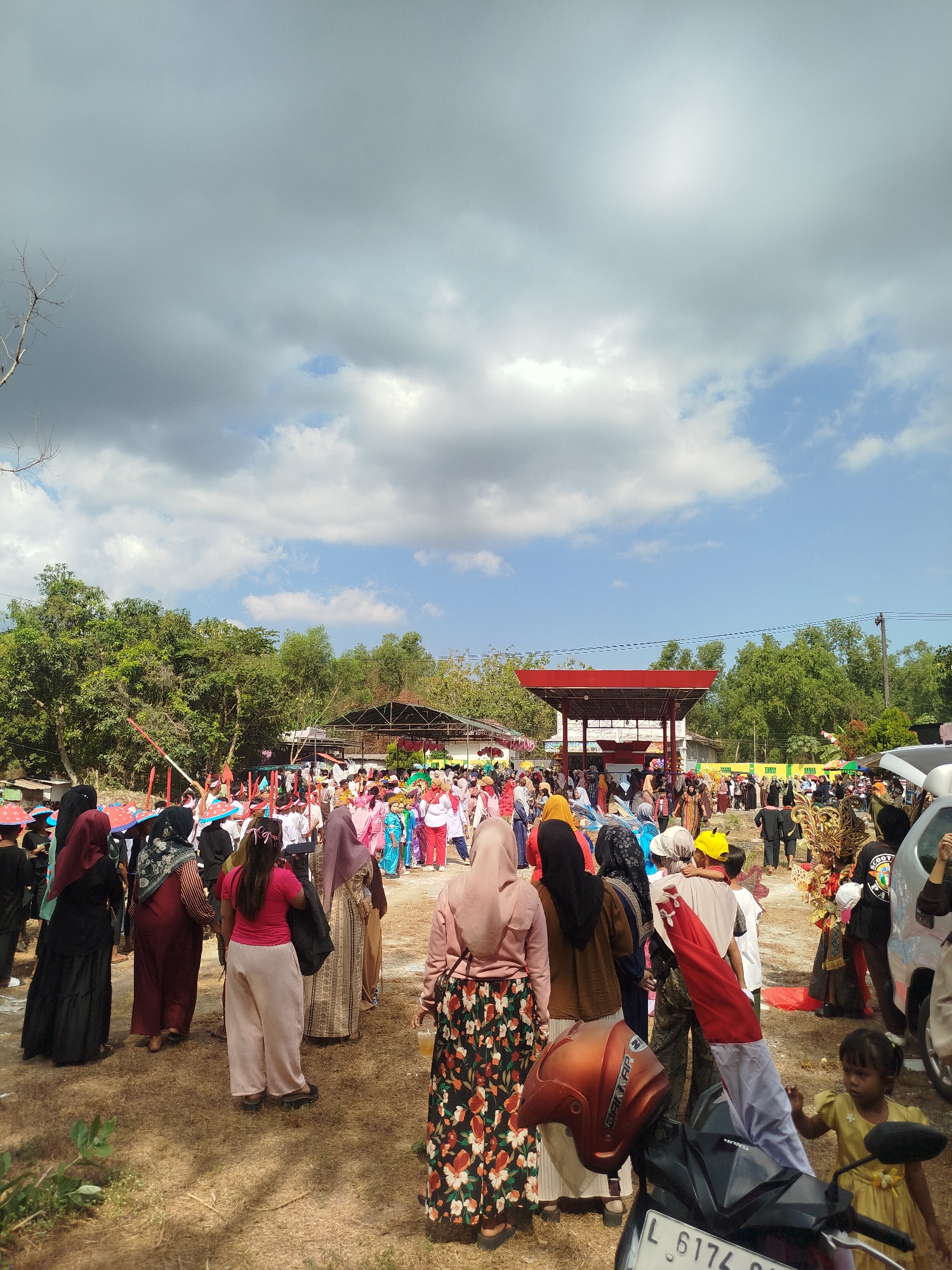 An outdoor daytime scene depicts a gathering of people, likely for a cultural event or festival, in Jambu, Indonesia. The foreground is dominated by dry, patchy brown grass, with a portion of a black and silver scooter visible, featuring a brown helmet and the Indonesian flag draped over it. A young girl in a yellow dress stands to the right of the scooter, looking towards the right. In the middle ground, a diverse crowd of people, many wearing traditional attire and headscarves, are observing an event in front of a red, open-sided structure with a canopy. Some individuals are holding colorful umbrellas or hats. Behind the structure, lush green trees form a backdrop. The sky above is a mix of bright blue patches and grey clouds. The overall atmosphere suggests a community event taking place on a sunny but partly cloudy day.