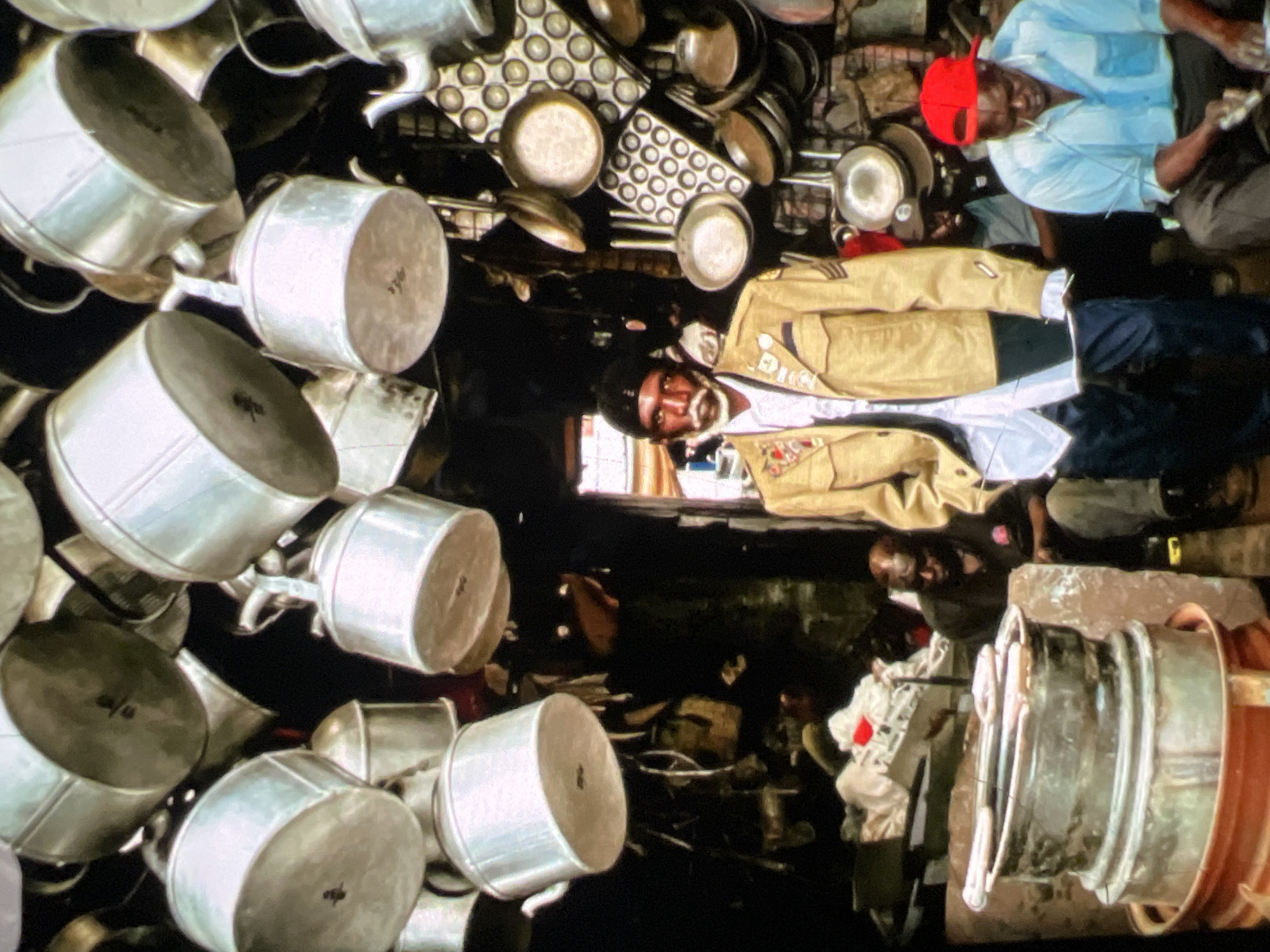 The image depicts a bustling, cluttered indoor market stall, likely in Lagos, Nigeria, characterized by an abundance of metal cookware, predominantly aluminum pots and pans, hanging from the ceiling and shelves. The stall appears to be a vendor of kitchenware. In the foreground, three men are visible. The central figure is a man with a mustache and beard, wearing a light-colored jacket adorned with badges and patches over a white shirt and dark pants. To his right, another man, wearing a light blue shirt and a red baseball cap, is seated and appears to be engaged in an activity, possibly preparing or examining something. Behind them, partially obscured, other individuals can be seen amongst the merchandise. The overall impression is one of a densely packed, active marketplace environment.
