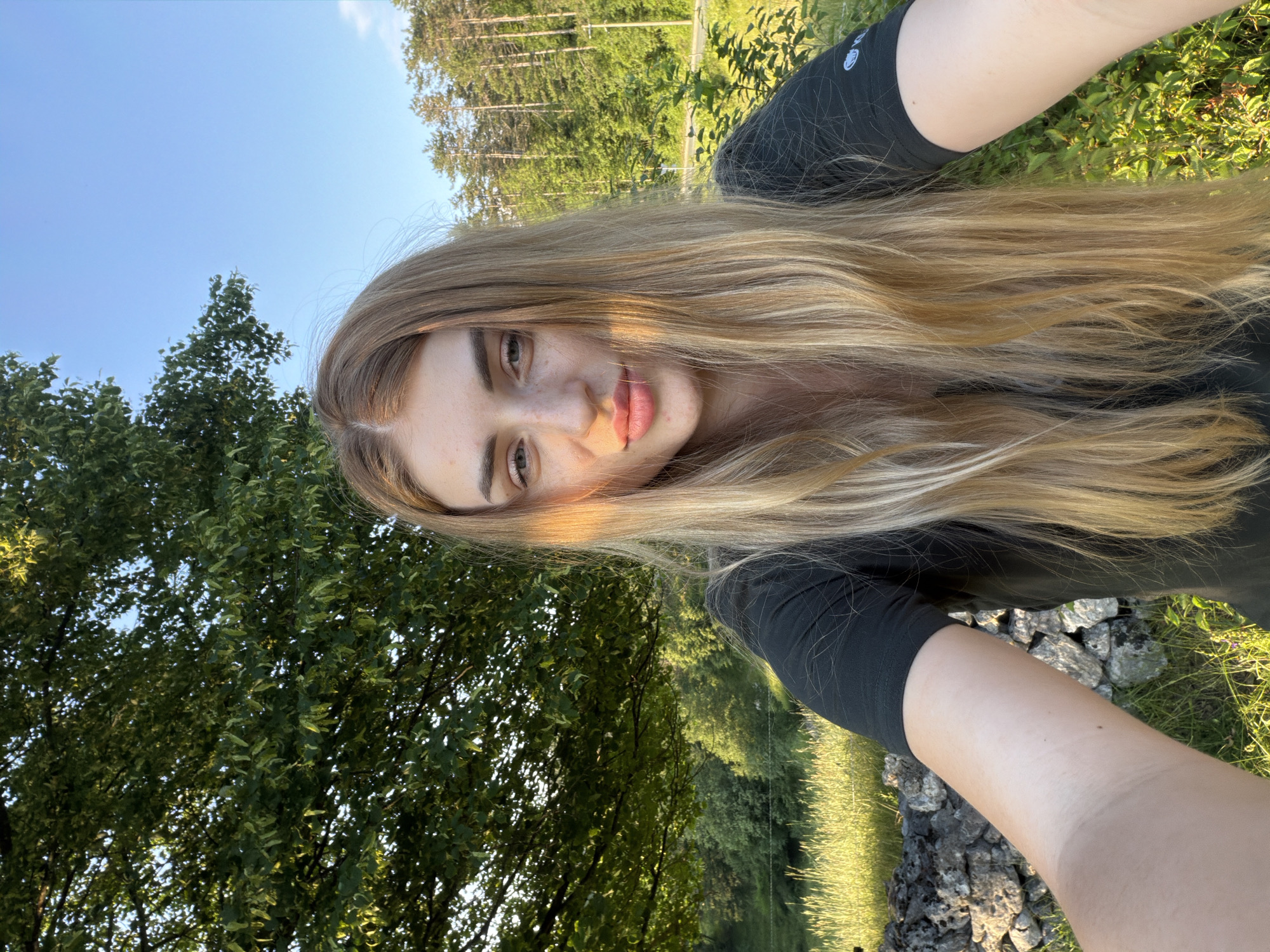 An outdoor, waist-up, selfie-style photograph shows a young, Caucasian woman with long, blonde hair, fair skin, and light blue eyes. She is wearing a black t-shirt and looking directly at the camera. Her head is tilted slightly to her left, and a streak of sunlight illuminates the left side of her face. She has a light dusting of freckles across her nose and cheeks. Her lips are slightly parted, and her eyebrows are well-defined.
The background is a natural landscape. To the left, there is a large, leafy green tree. To the right and behind her, a line of tall, slender pine trees stretches across the frame, with a bright blue sky visible above. In the middle ground, there is a grassy field with a low stone wall in the foreground on the left. The lighting suggests it is late afternoon or early evening, with the sun casting long shadows and warm light. The location is identified as Unknown, Unknown.