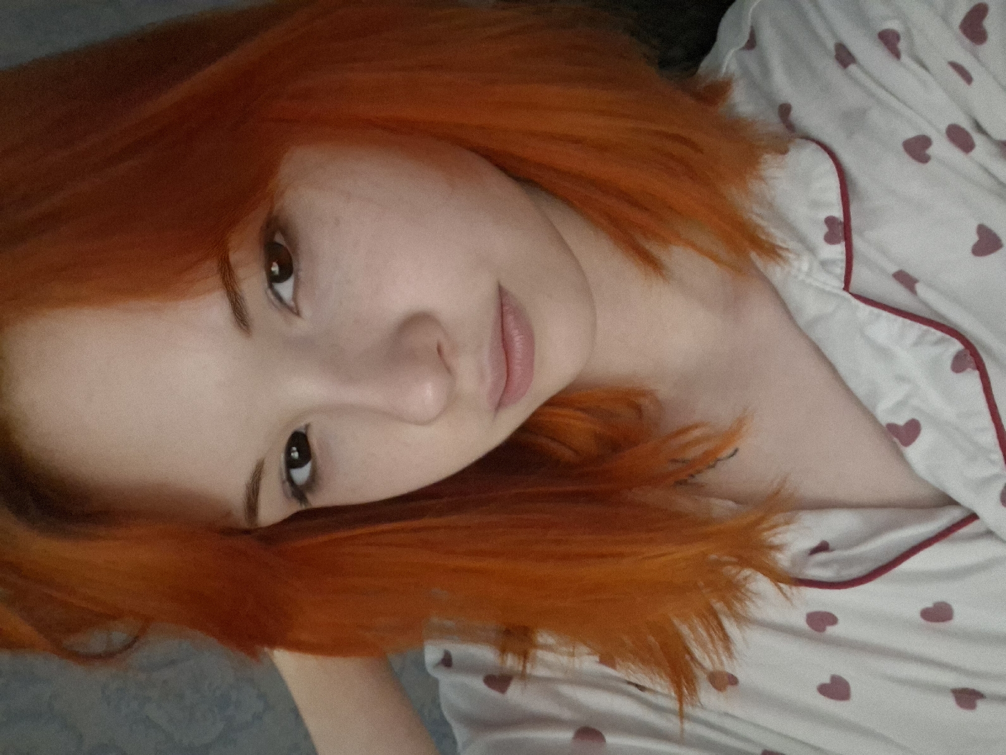 A close-up shot shows a fair-skinned person with bright orange, shoulder-length hair, appearing to lie down. The person has dark eyes and a neutral expression. They are wearing a white top or pajamas patterned with small red hearts and trimmed with red piping. The background is slightly blurred, showing a light-colored surface. It is not possible to confirm the location of the picture from the provided information.